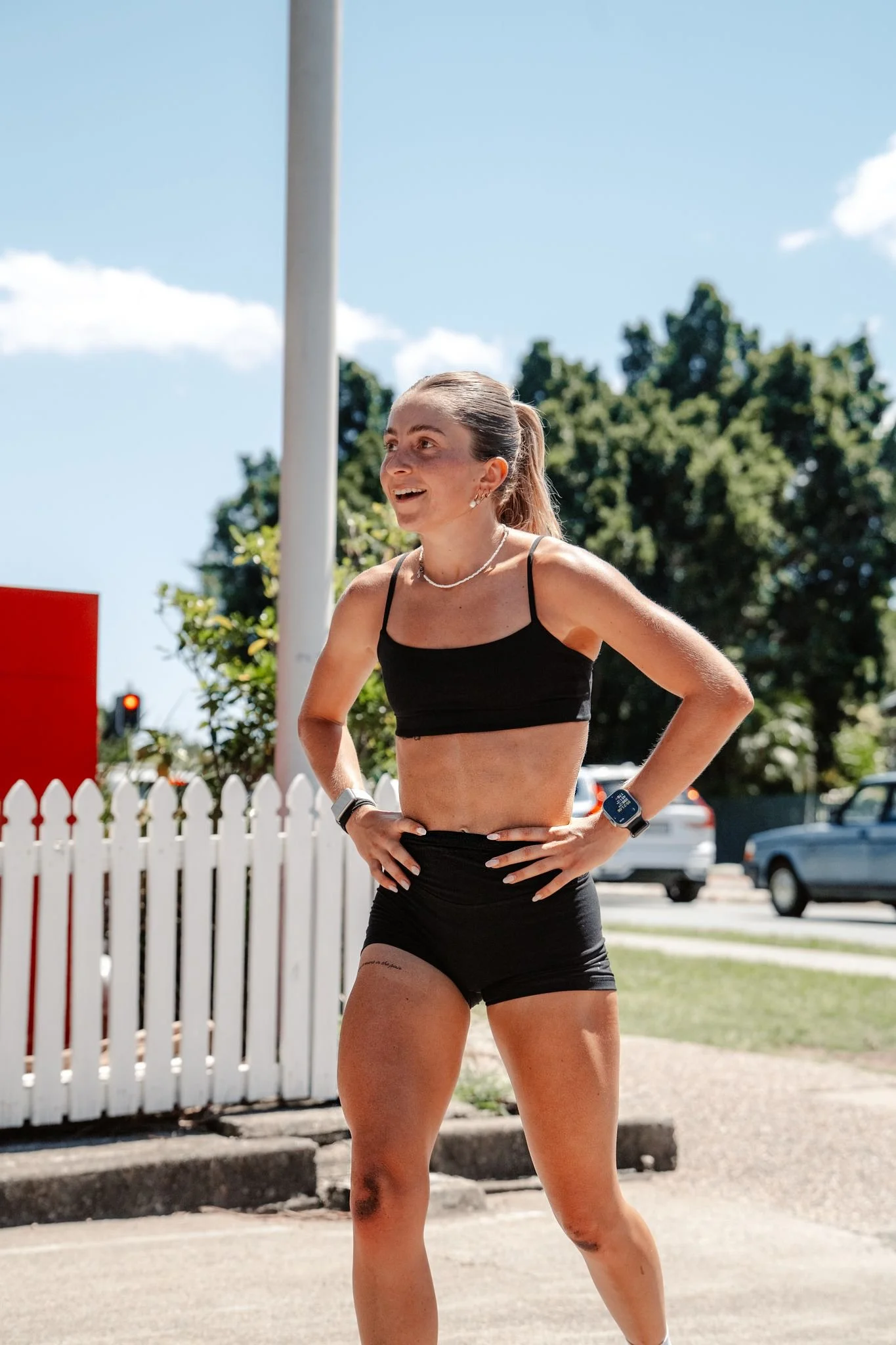 A woman running outdoors, wearing black athletic clothing, a watch, and a necklace, smiling with her hands on her hips against a background of blue sky, trees, cars, and a white fence.