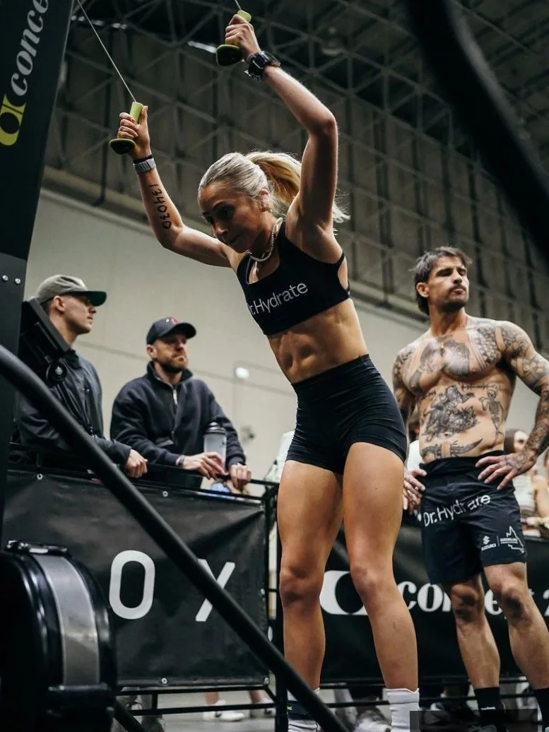 A woman with blonde hair in a ponytail wearing black workout clothes, lifting dumbbells overhead in a gym. Two men with tattoos and wearing black shorts and t-shirts stand nearby. Kids are in the background, and gym equipment and banners are visible.