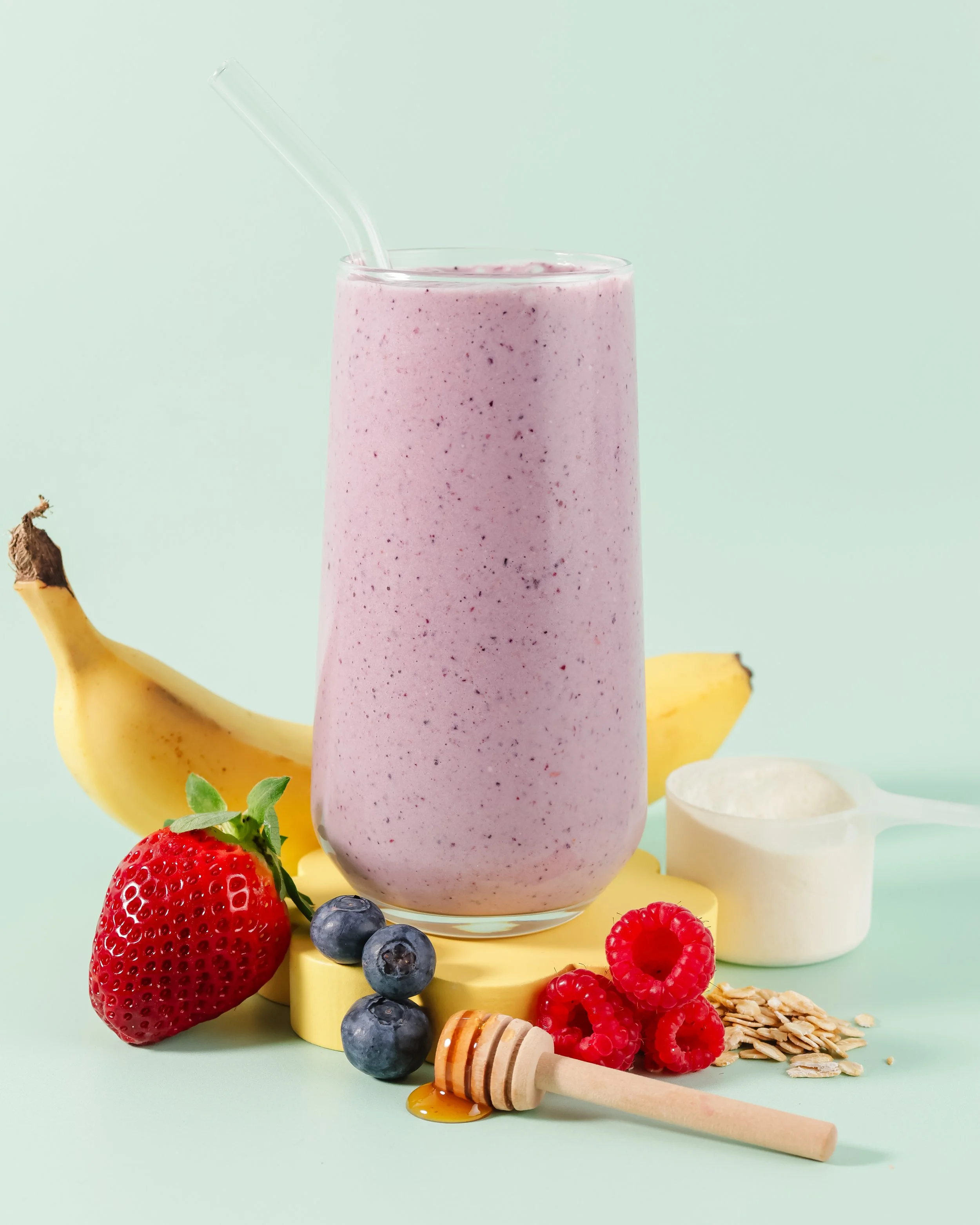 A pink berry smoothie in a tall glass with a straw, surrounded by fresh strawberries, blueberries, raspberries, oatmeal, honey, and a honey dipper on a pastel background.