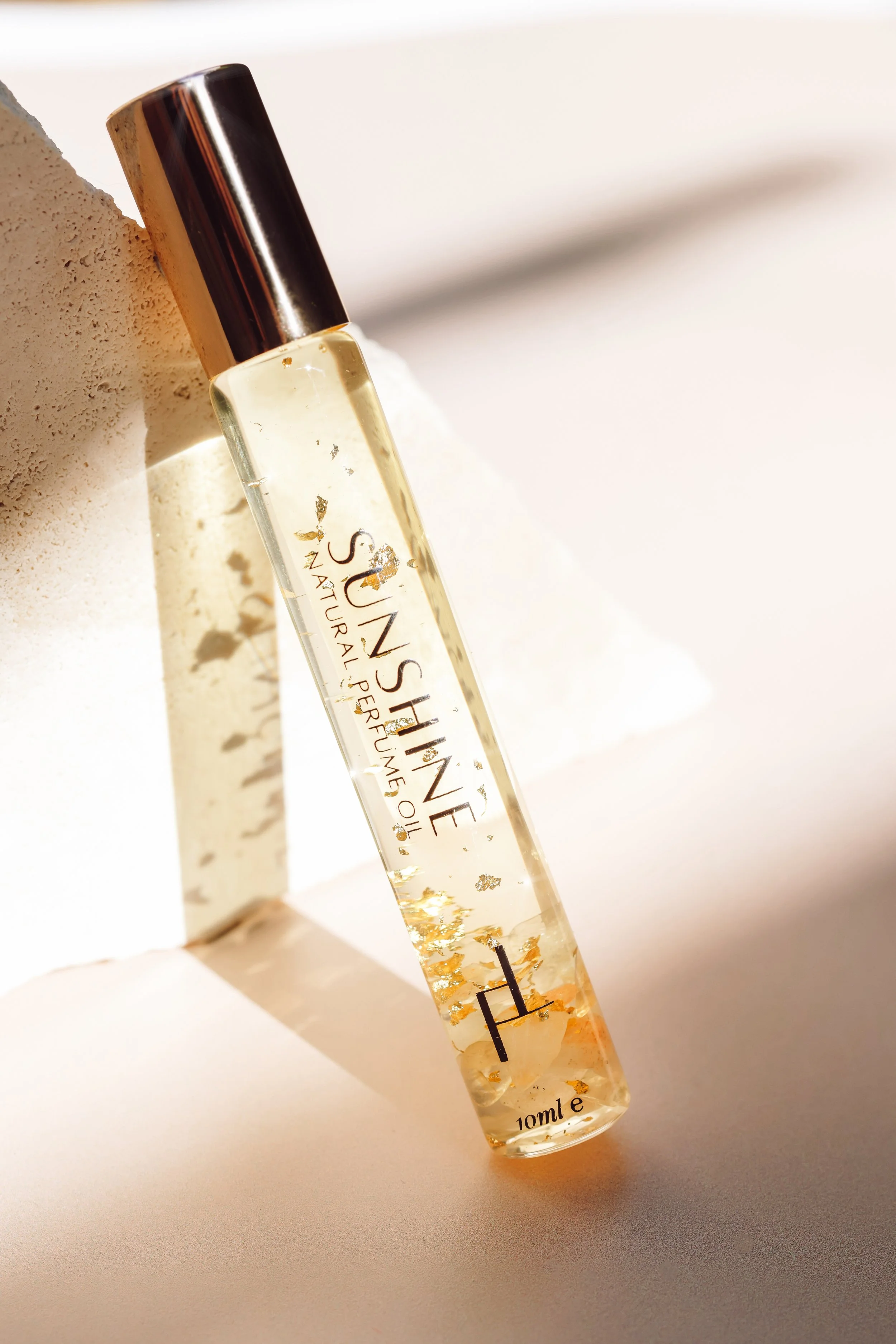 A clear glass perfume rollerball with a black cap, containing a light-colored liquid with gold flakes, resting on a light surface with shadows.