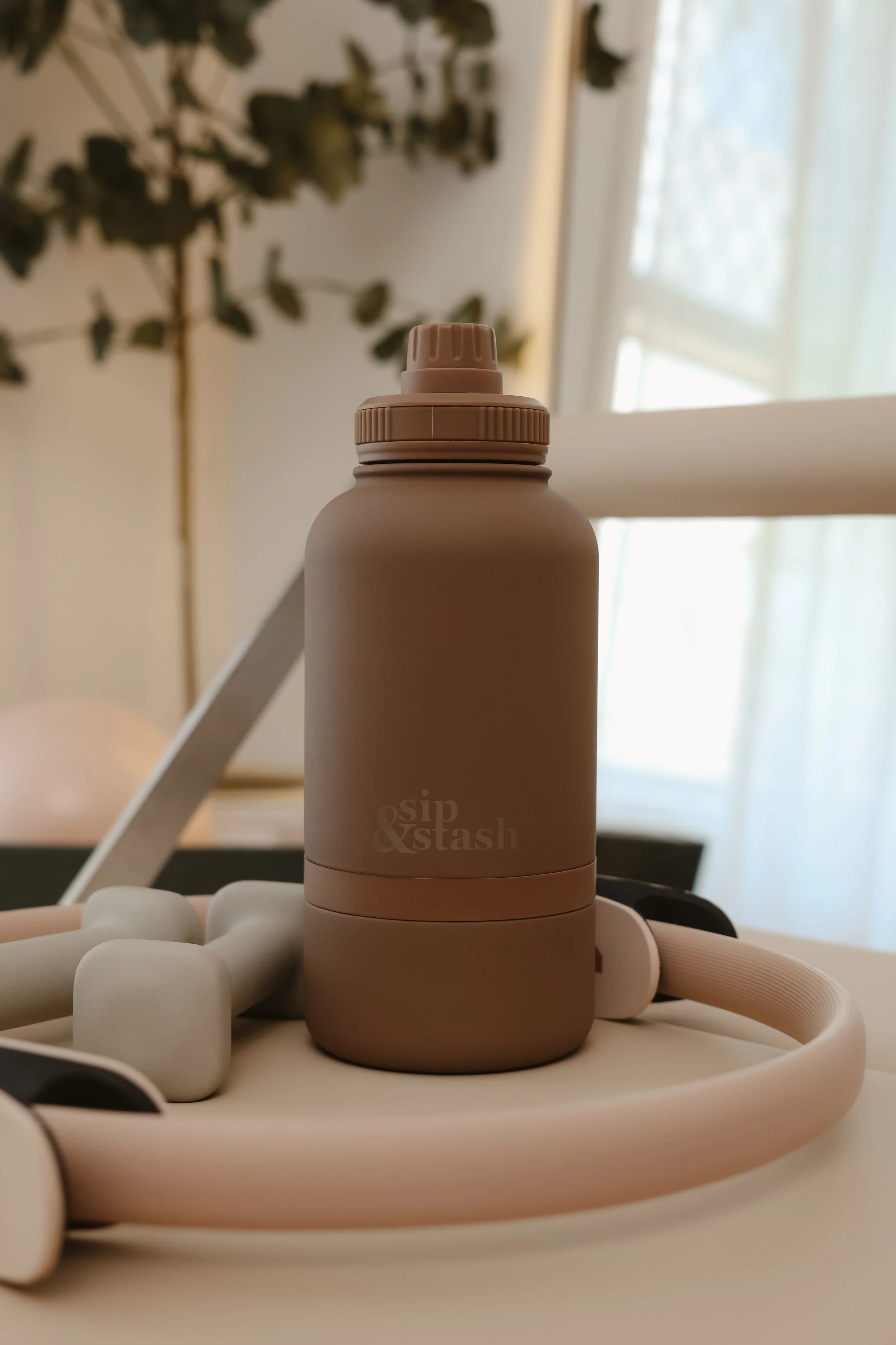 A beige bottle with a white cap sitting on a white tray with white rounded objects and flexible tubes, with a blurred background of a window with natural light.