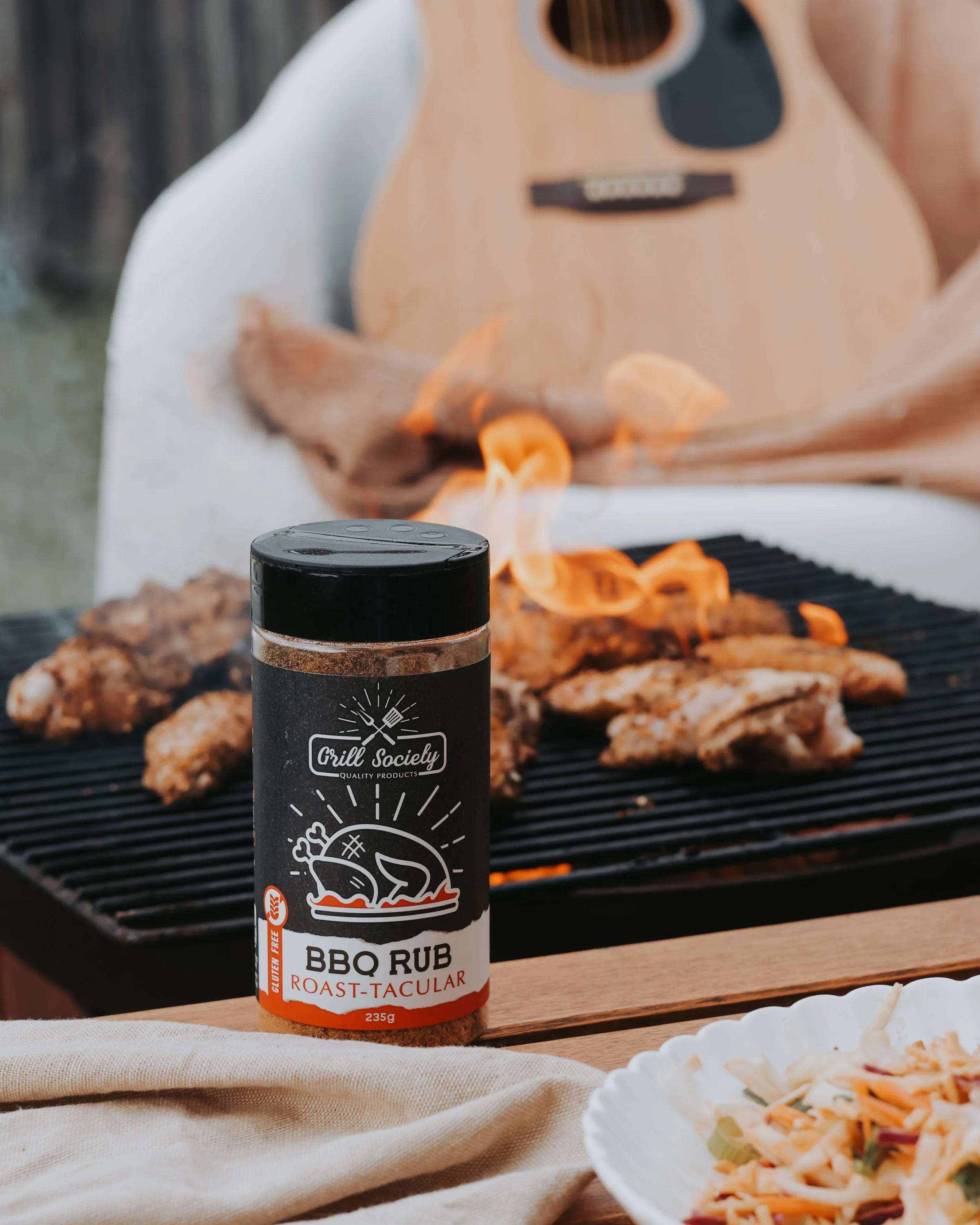Bottle of grill BBQ rub on a table in front of a grill with flaming food, with a plate of pasta salad in the foreground