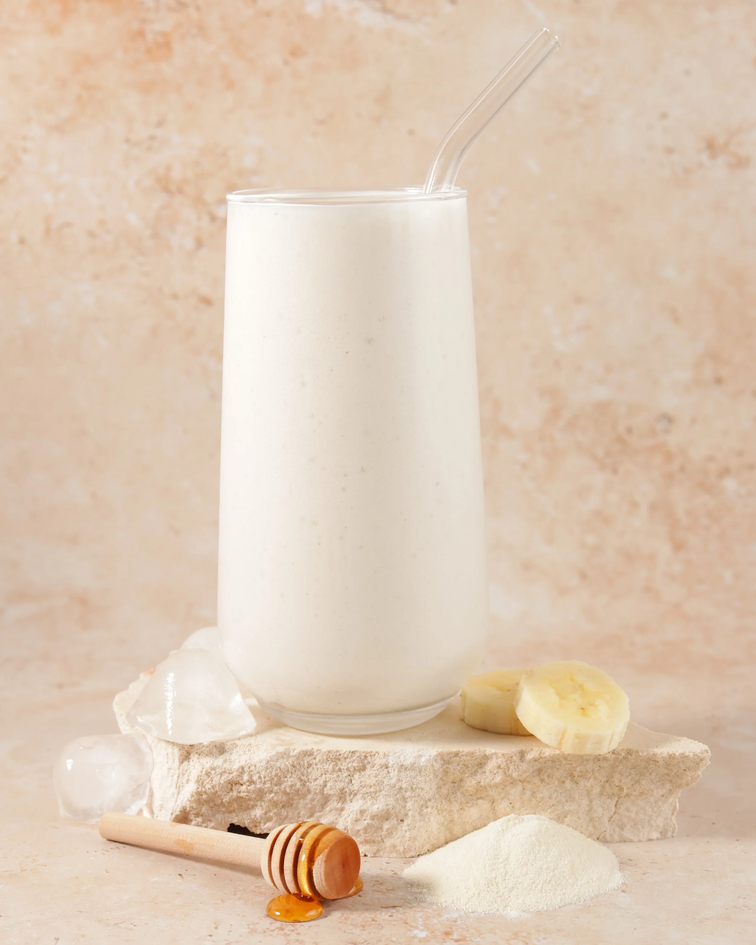 A glass of milk with a clear straw, surrounded by honey, flour, and chunks of butter on a textured surface with a neutral background.