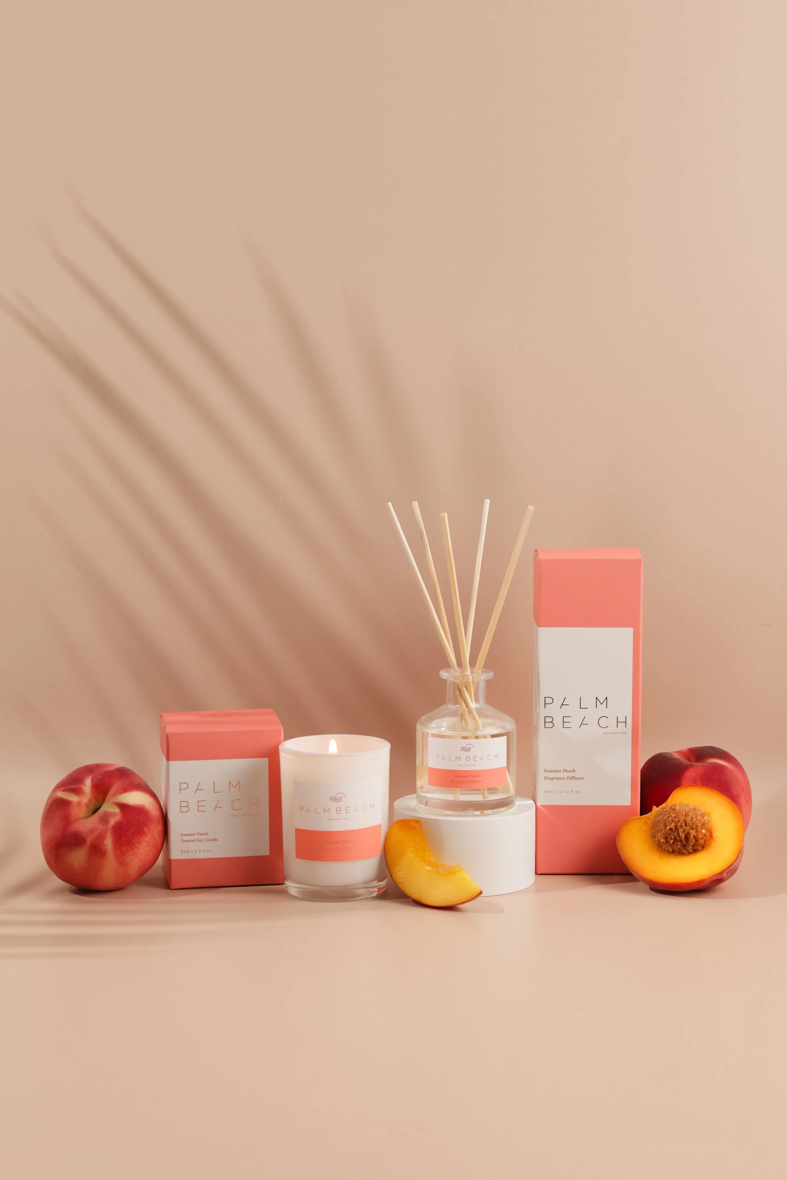 Arrangement of peach-themed Palm Beach scented candle and fragrance diffuser boxes, peaches, apple, and a candle holder with reed sticks against a blush-colored wall with shadow.