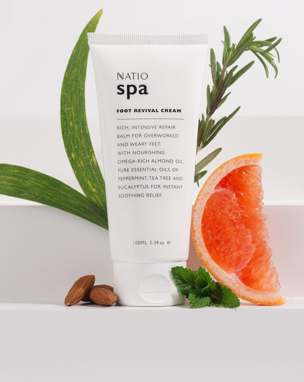 A white tube of Natio Spa Foot Revival Cream with ingredients listed, surrounded by grapefruit, rosemary, mint, aloe vera leaves, and almonds on a white surface.