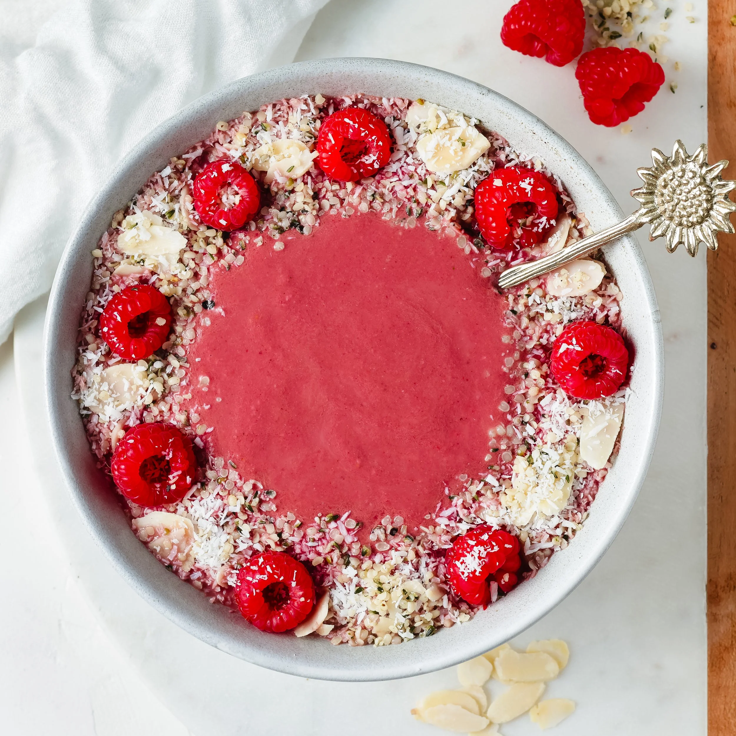 A bowl of pink smoothie decorated with raspberries, white chocolate shavings, coconut flakes, and chopped nuts with more raspberries on a white surface.