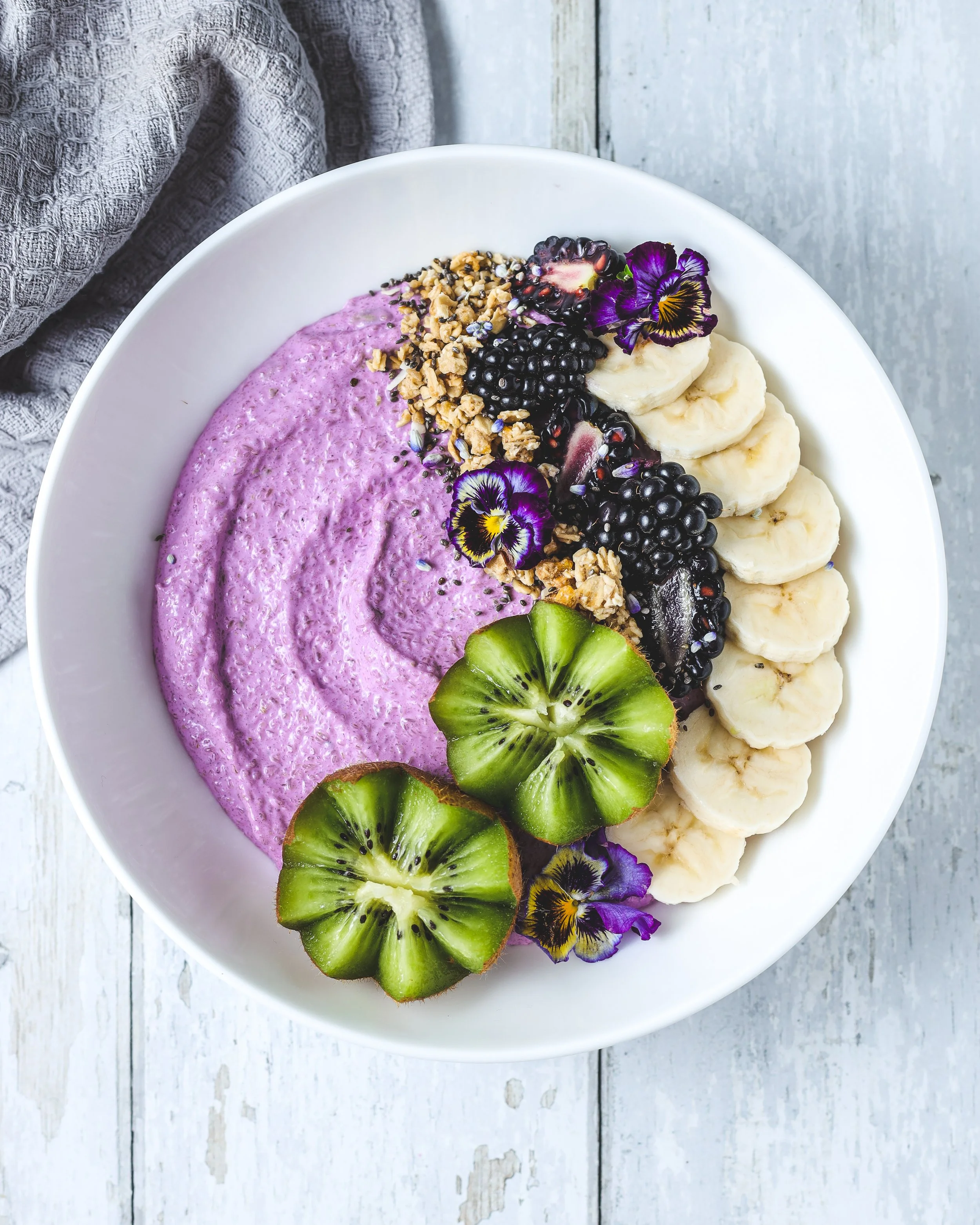 A white bowl of purple smoothie garnished with sliced banana, kiwi, blackberries, purple edible flowers, granola, and seeds on a white wooden surface.