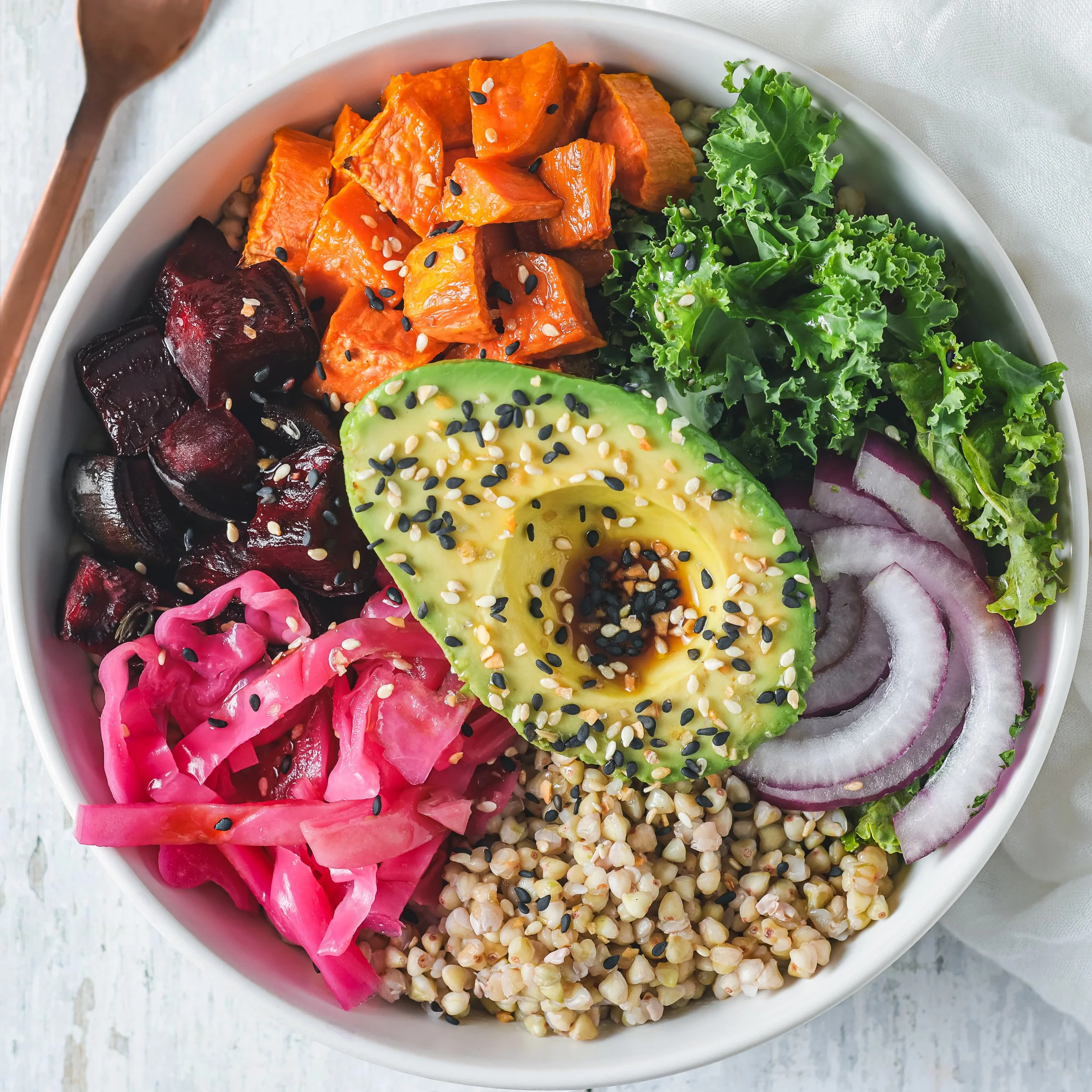 A white bowl containing a salad with roasted sweet potatoes topped with black and white sesame seeds, sliced avocado with sesame seeds, beets, pickled red onions, shredded carrots, kale, and other vegetables on a white wooden surface.