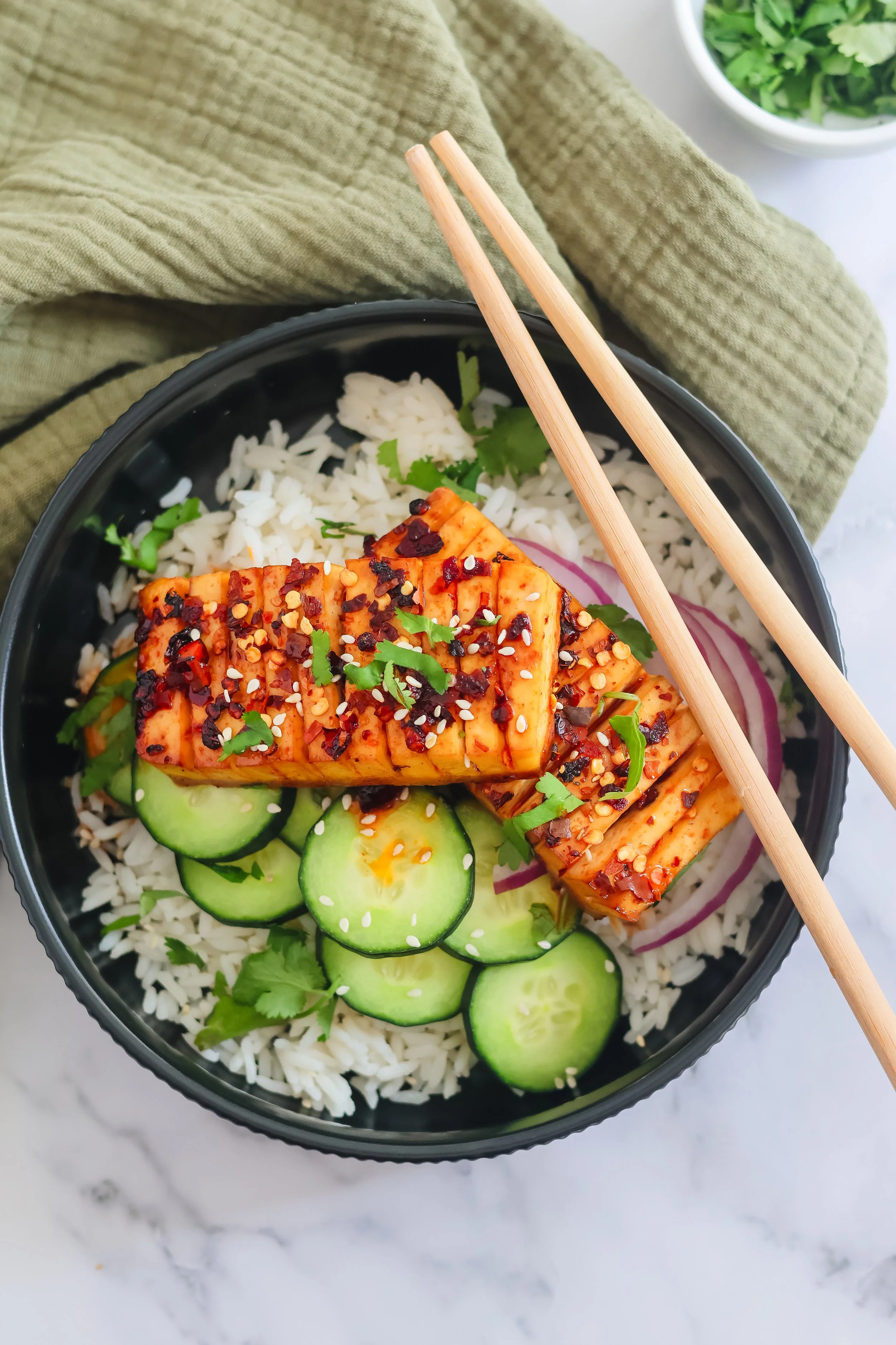A bowl of rice topped with sliced cucumbers, red onion, cilantro, and spicy marinated tofu garnished with chili flakes and sesame seeds, with a side of greens in a white bowl.
