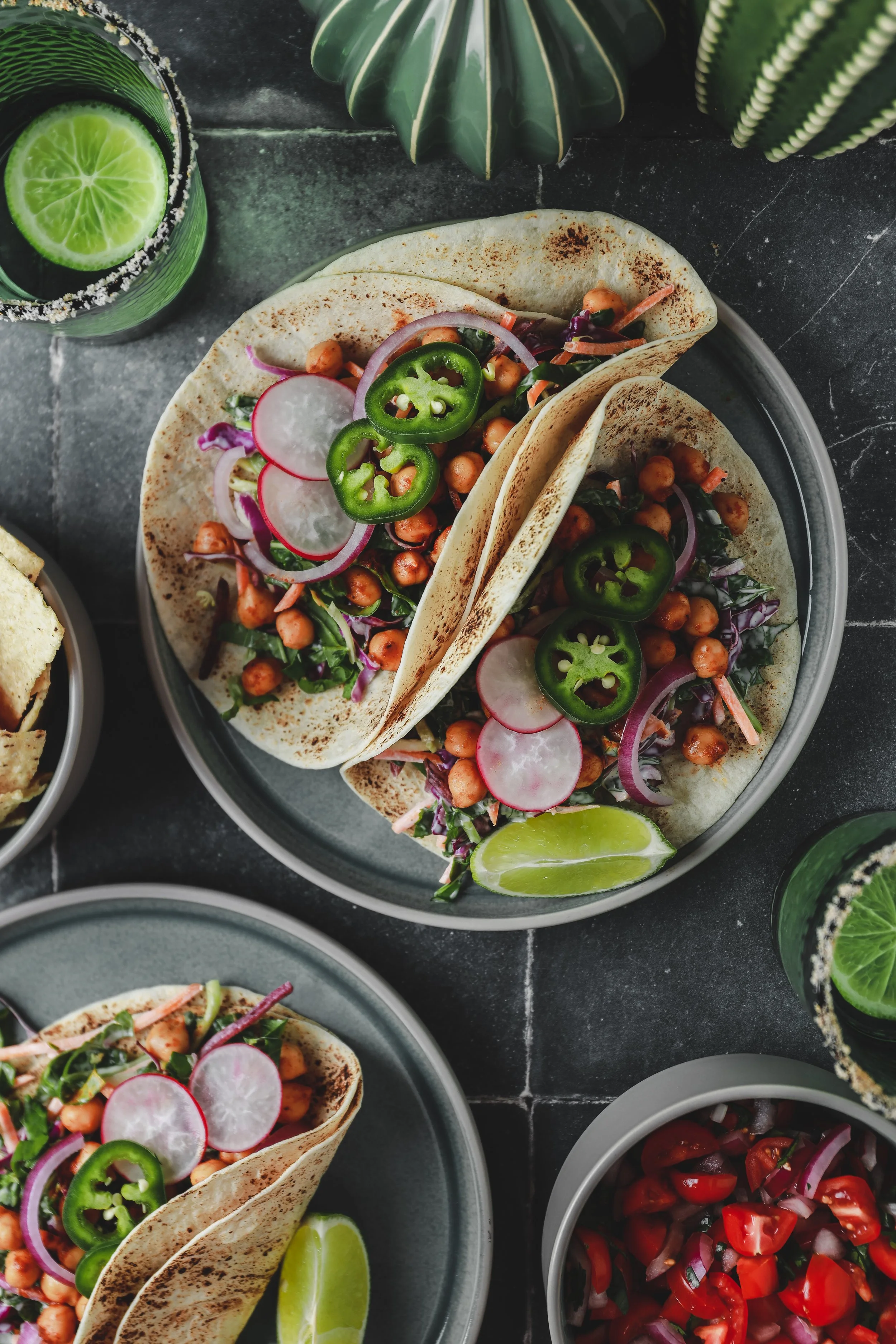 Close-up of two tacos filled with chickpeas, sliced radishes, onion, and jalapeño peppers, garnished with lime wedges on a dark surface.