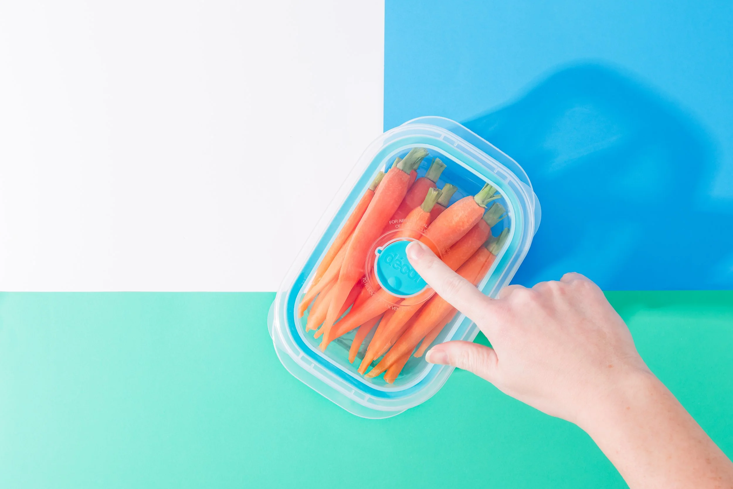 A hand holding a clear plastic container filled with fresh orange carrots against a colorful background of white, blue, and teal sections.