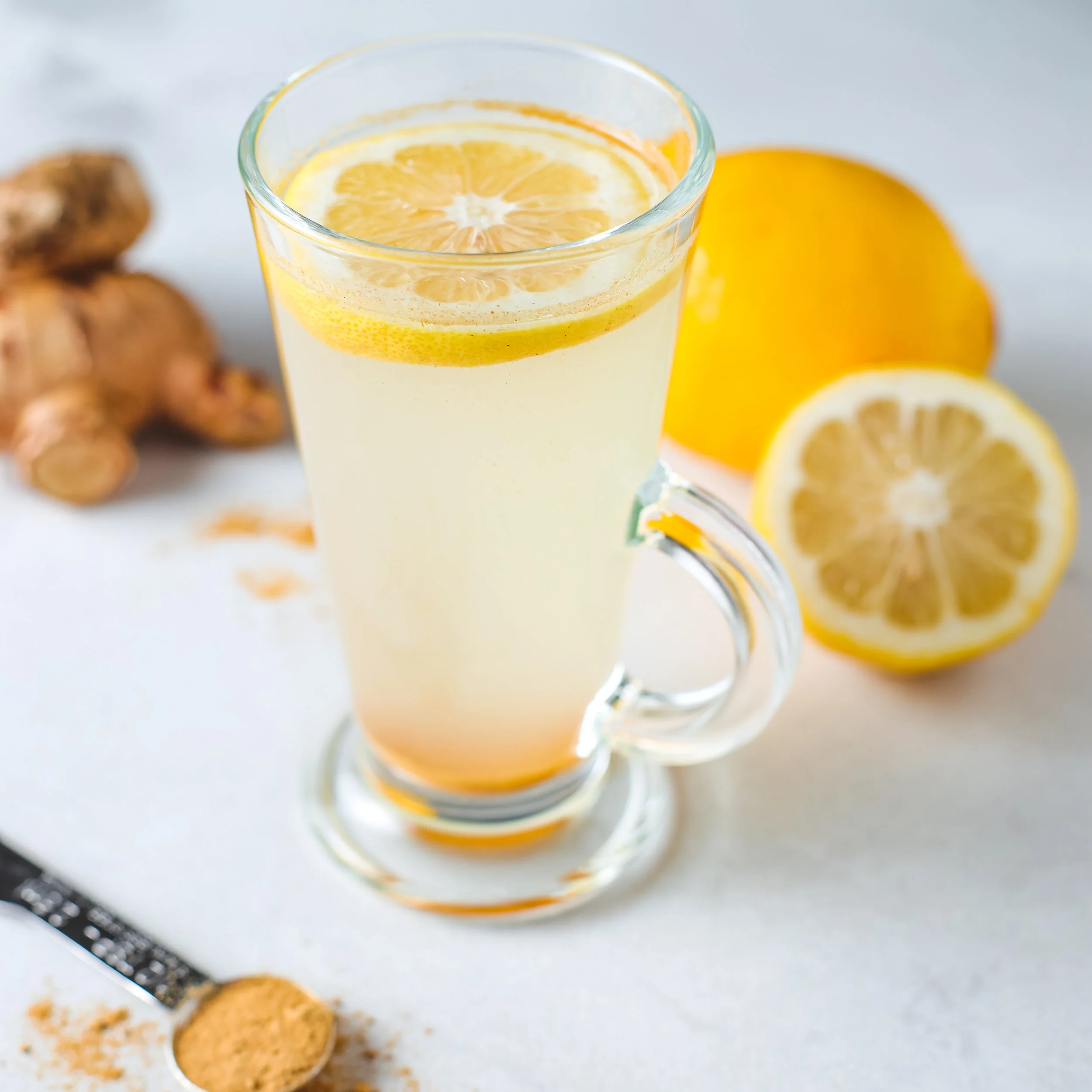 A glass of lemon-infused water with lemon slices, a lemon, ginger pieces, and cookies on a white surface.