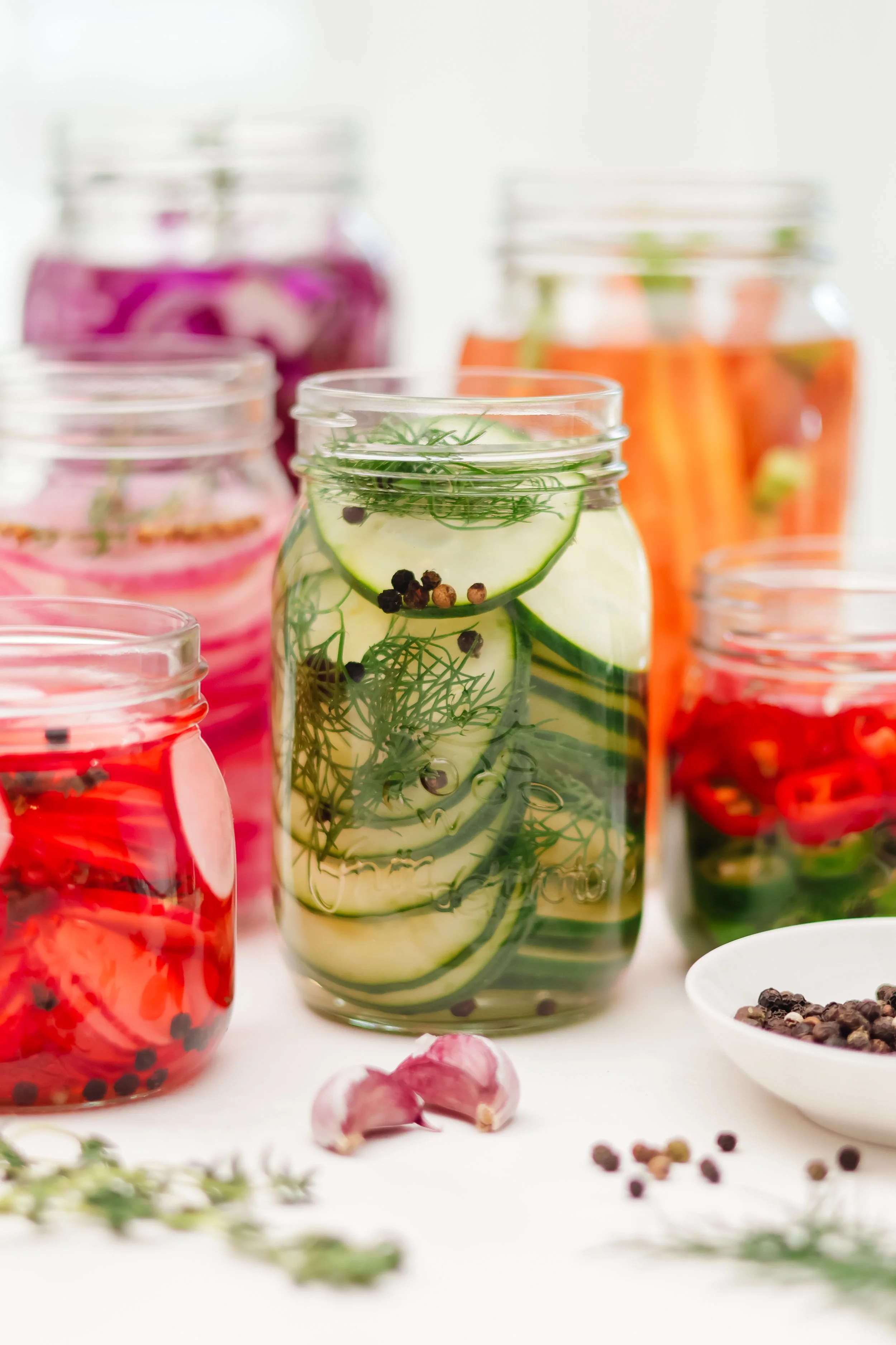 Several glass jars filled with colorful pickled vegetables and herbs, including cucumbers, carrots, and red onions, with scattered peppercorns and garlic cloves in a white background.