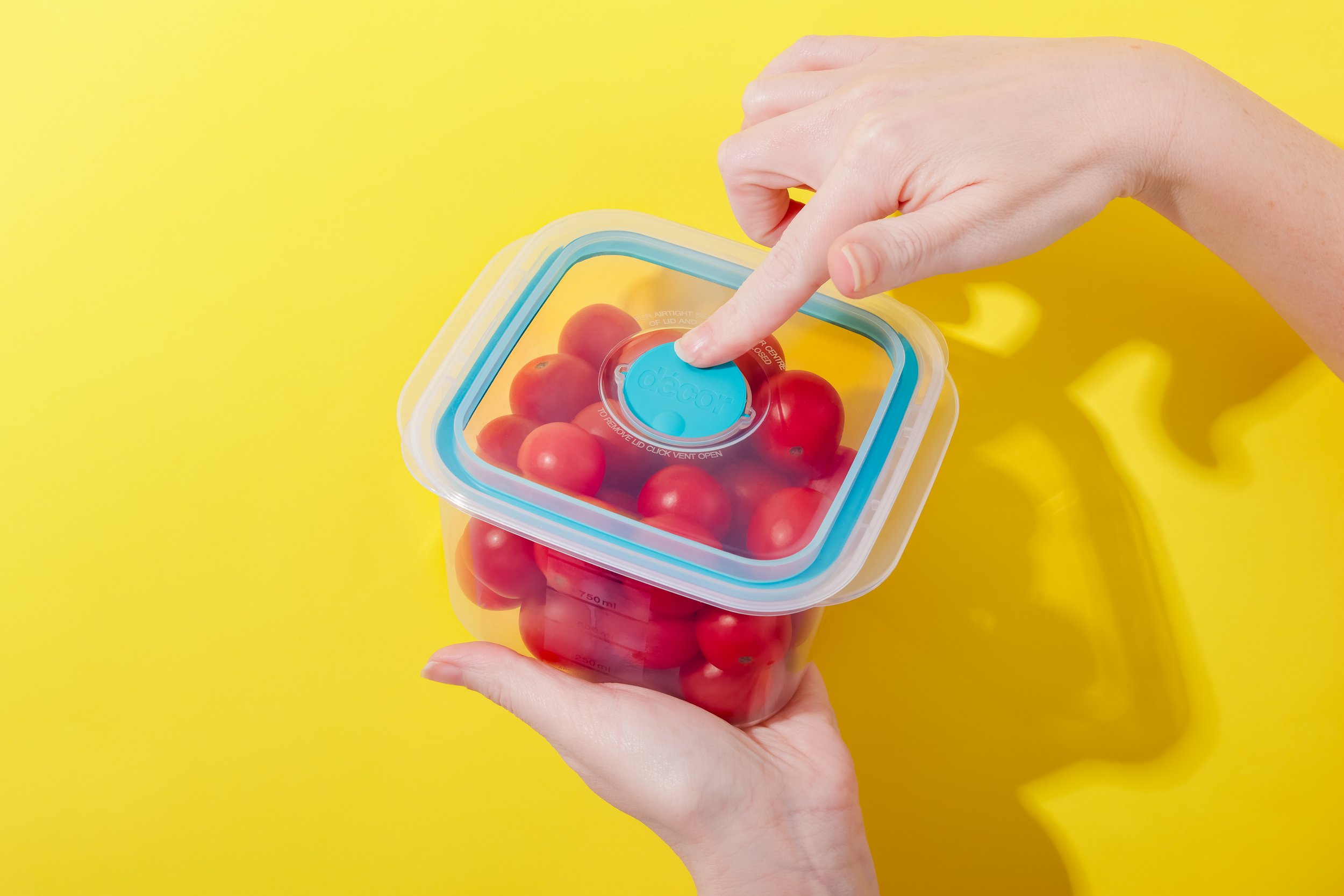 Person opening a plastic container filled with cherry tomatoes against a bright yellow background.
