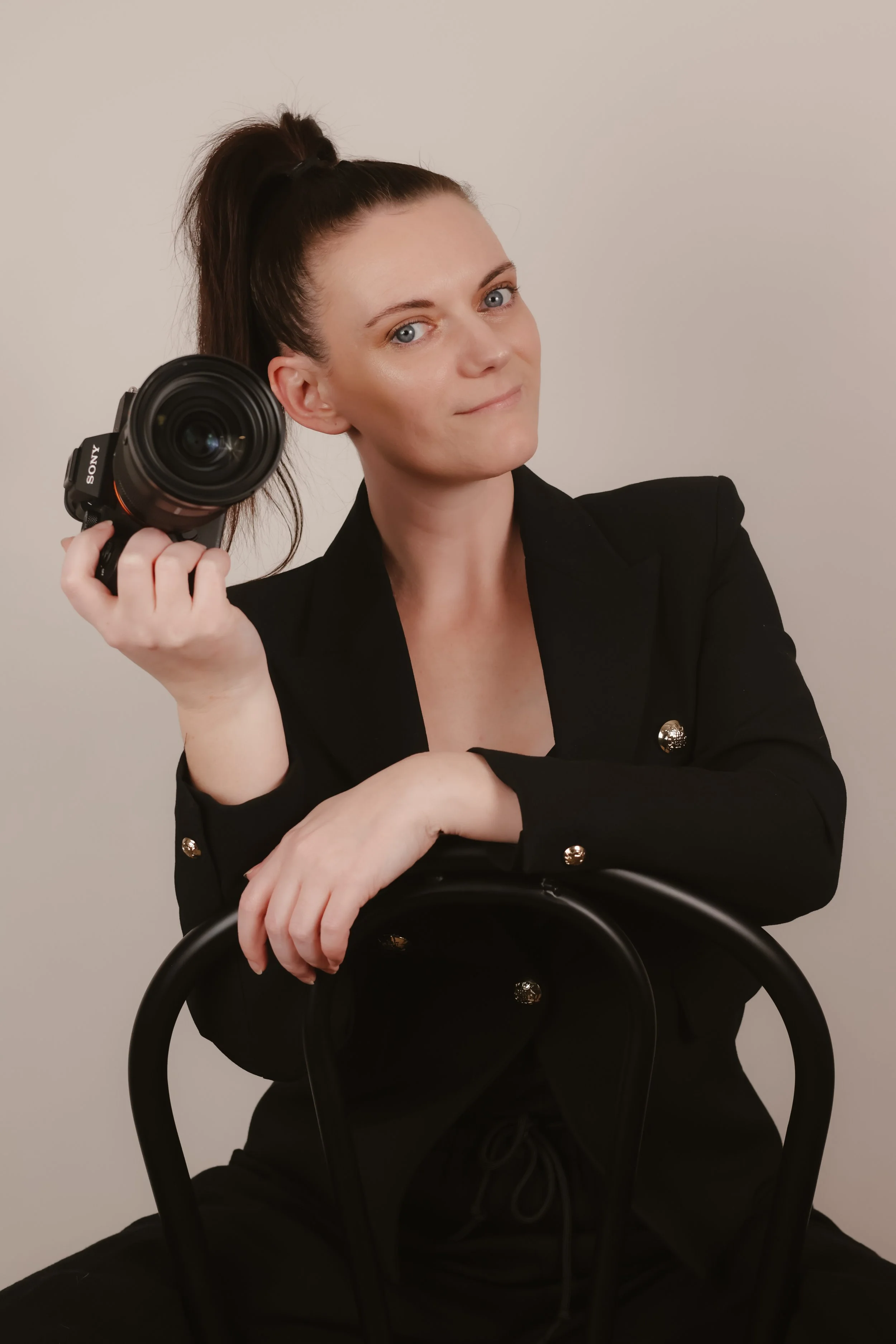 A woman with dark hair in a high ponytail, wearing a black blazer with gold buttons, sitting on a black chair, holding a camera in her right hand, and resting her left arm on the chair's back, against a plain white background.