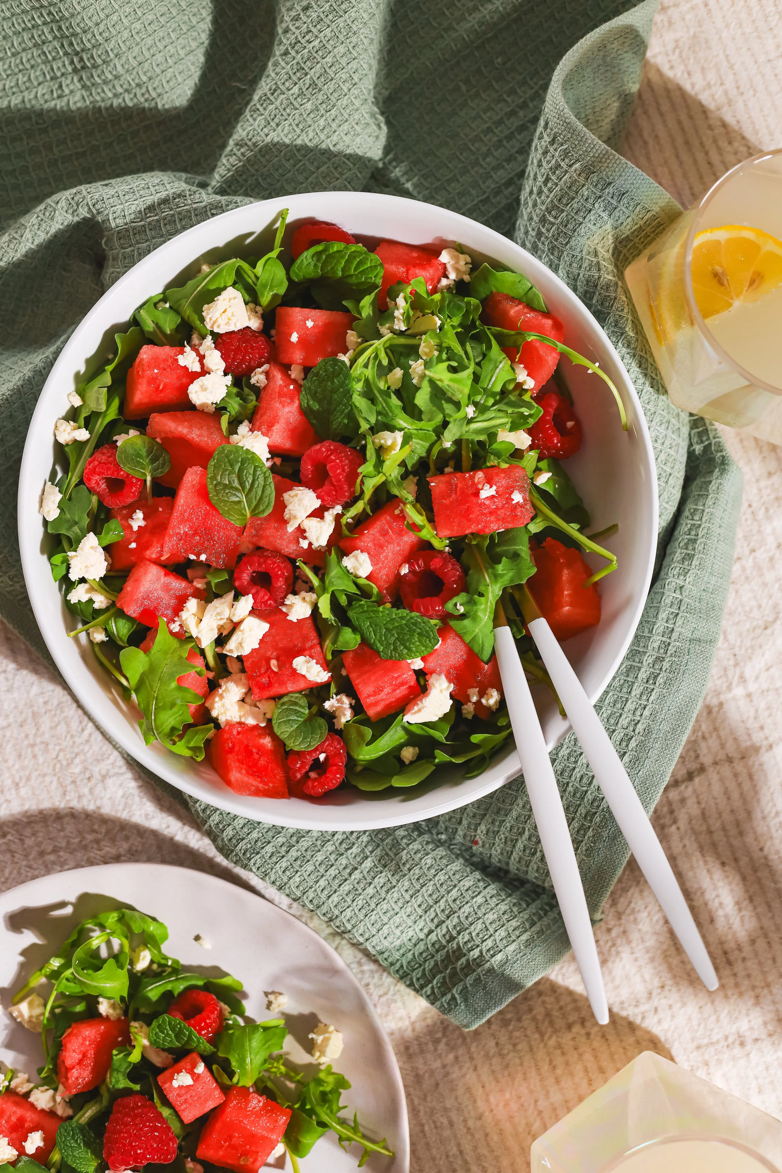 A bowl of fresh watermelon, raspberries, arugula, crumbled feta cheese, and mint leaves, with a glass of lemonade with lemon slices nearby, on a green cloth napkin on a beige table.