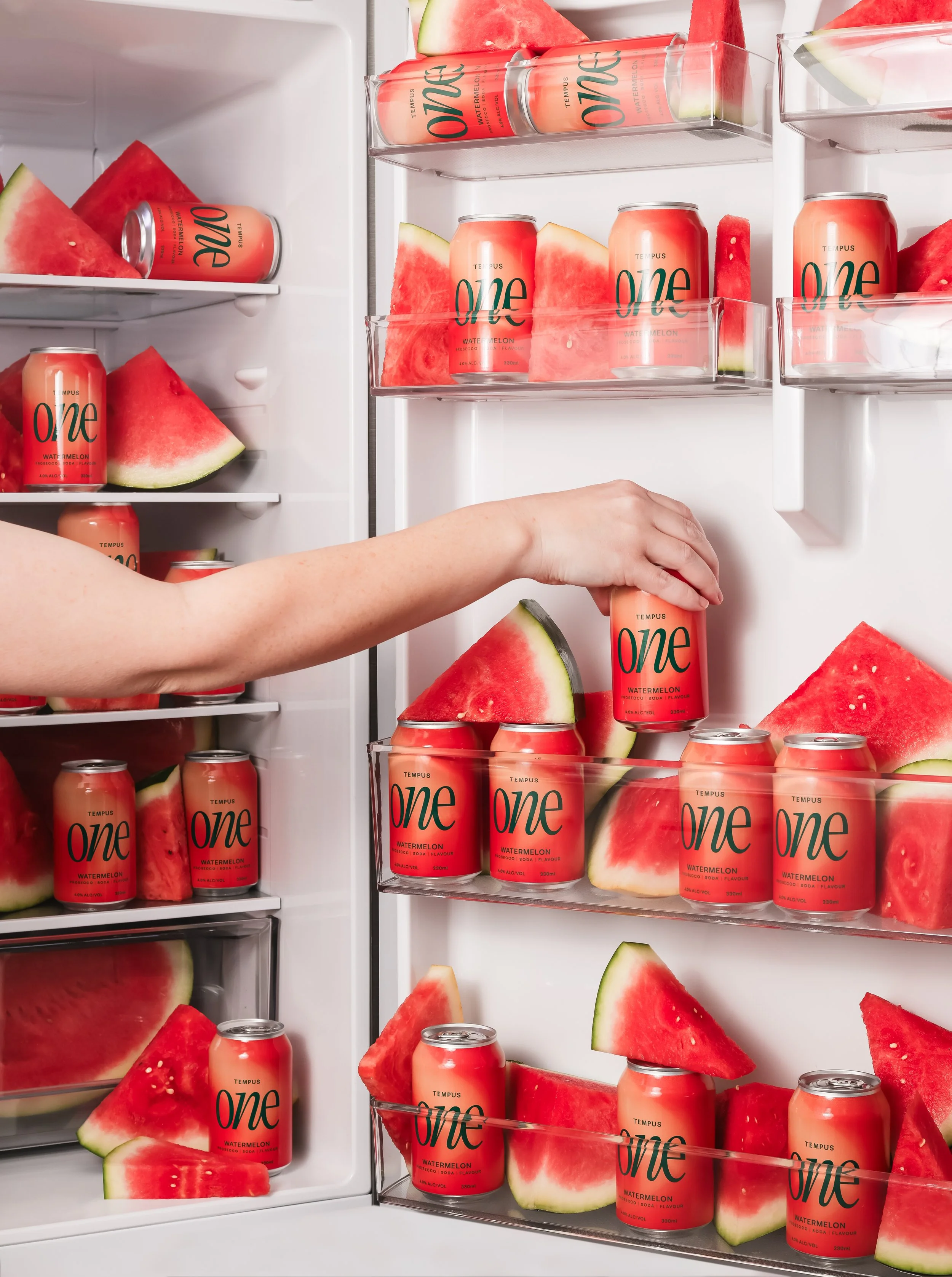 An open refrigerator door filled with cans of watermelon-flavored carbonated beverage called 'Tempus One Watermelon' and large slices of fresh watermelon. The cans are arranged on shelves and in door compartments, emphasizing the watermelon's red col