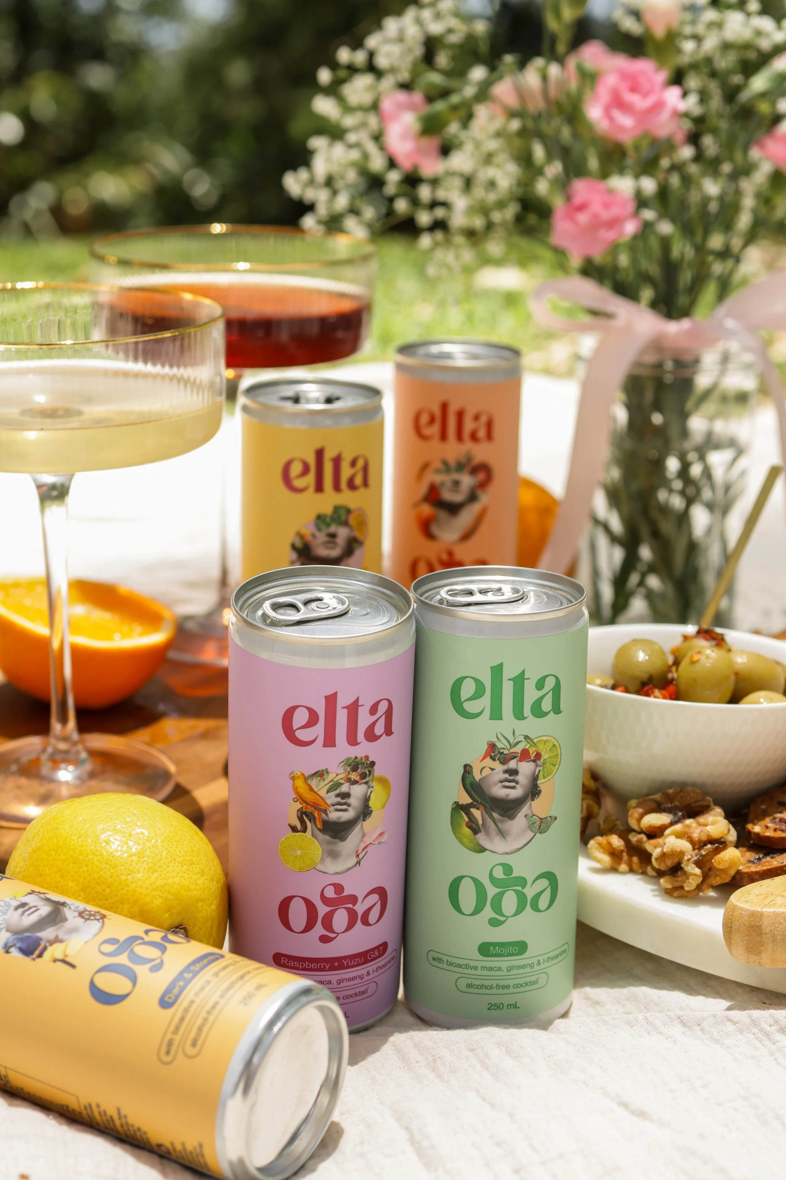 A table with colorful cans of Elta prebiotics drinks, glasses of wine, a bowl of olives, and a bouquet of pink and white flowers in a jar, outdoors on a sunny day.