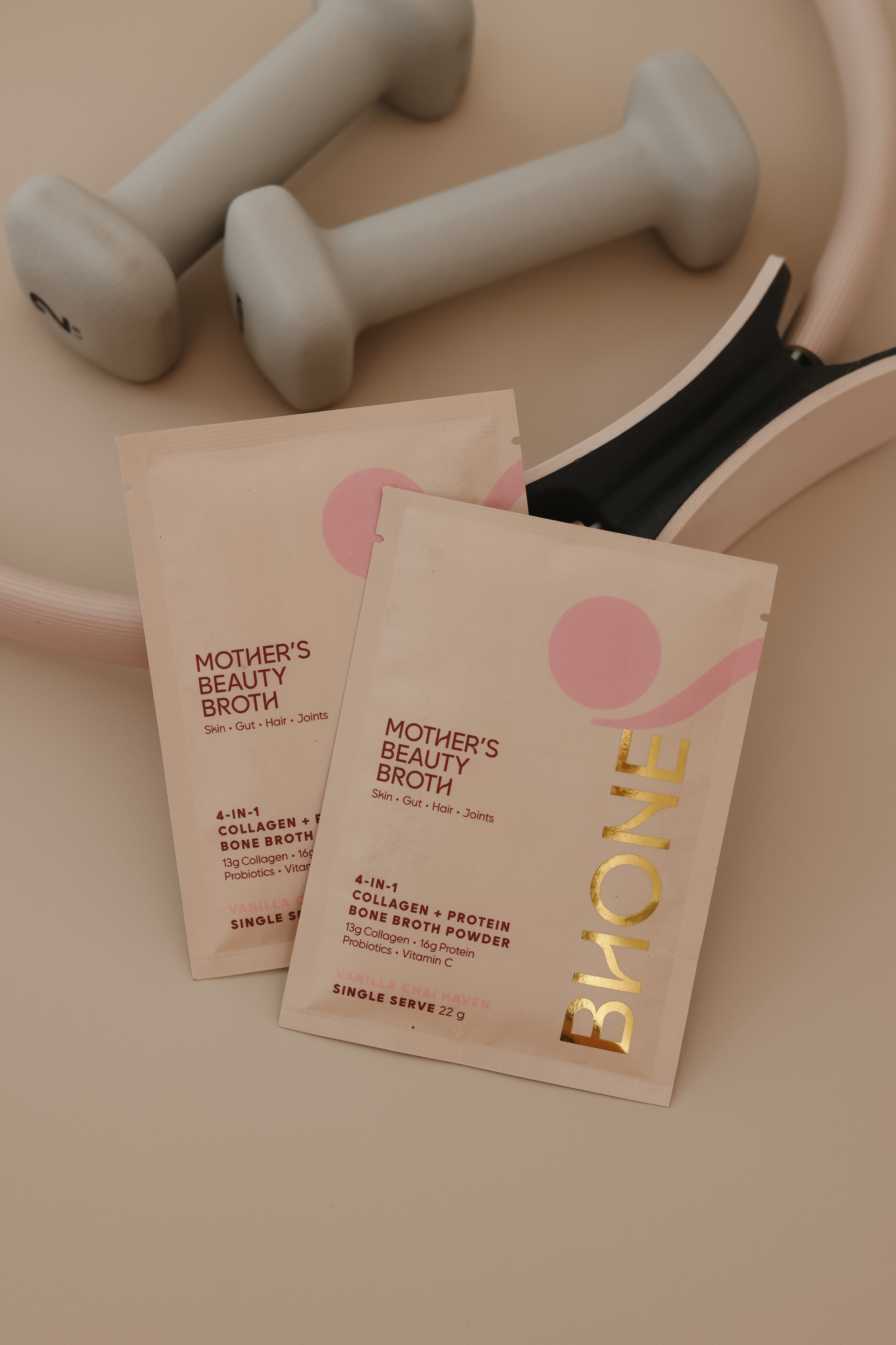 Close-up of two sample packets from Mother’s Beauty Broth line, labeled '4-in-1 Collagen + Protein Bone Broth Powder' with flavors vanilla chai haven and single serve. The packets are light pink with red and gold text.