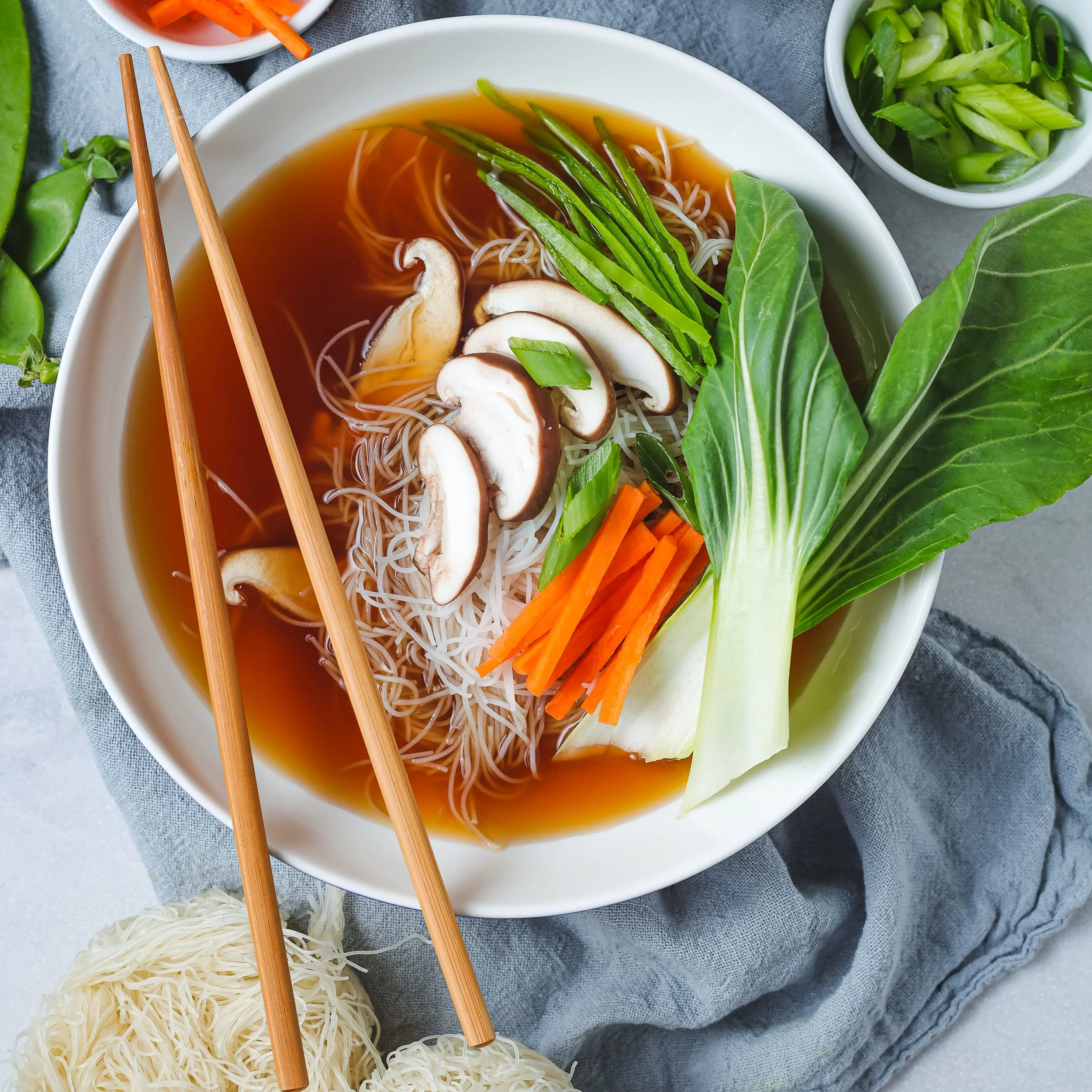 A bowl of Asian-style noodle soup with sliced mushrooms, chopped green onions, and shredded carrots, surrounded by bowls of chopped vegetables and rice noodles.