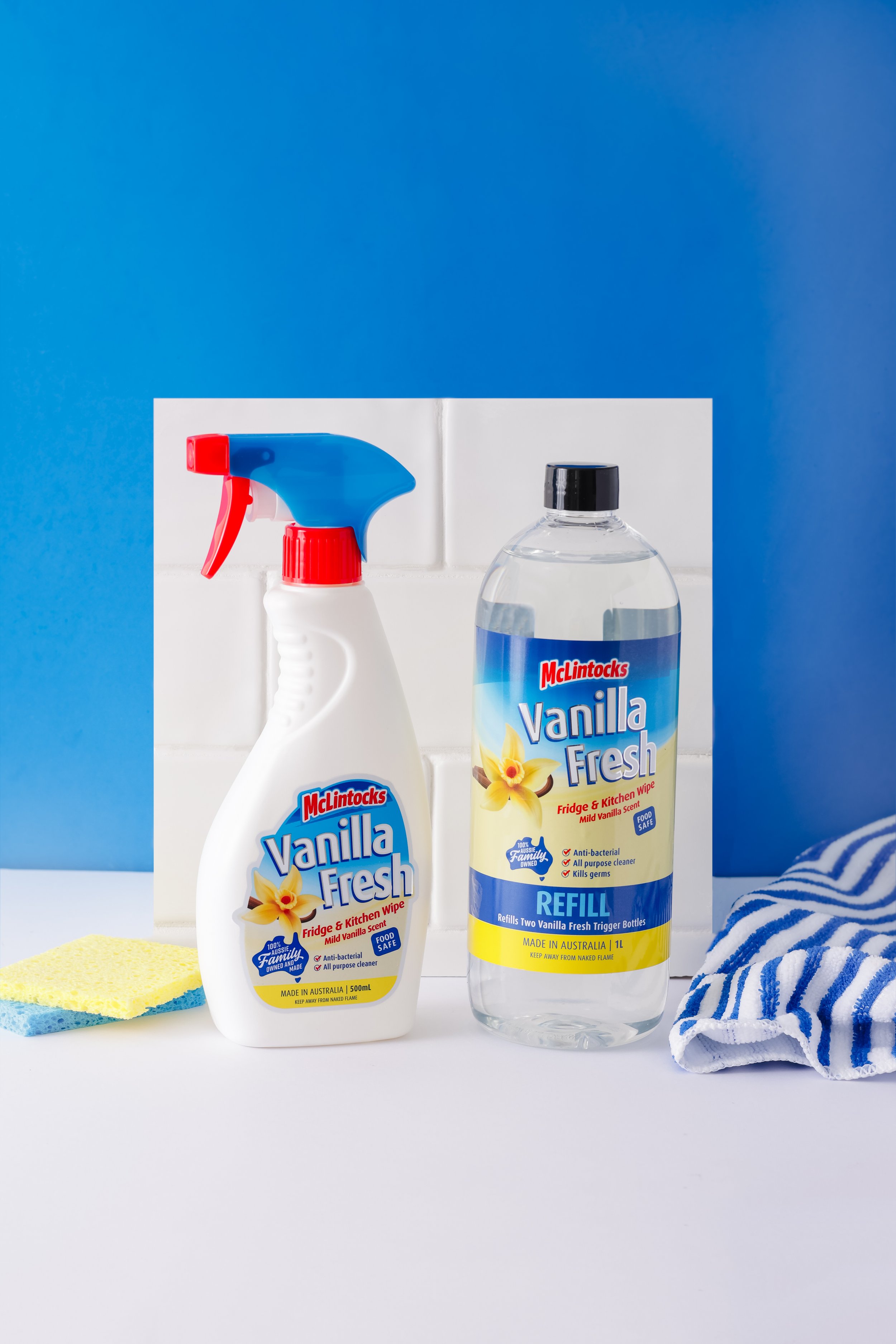 Cleaning supplies on a white surface with yellow and blue sponges and a striped cloth. In the background, there are two bottles of McLintocks Vanilla Fresh cleaner, one with a spray nozzle and the other a refill bottle.