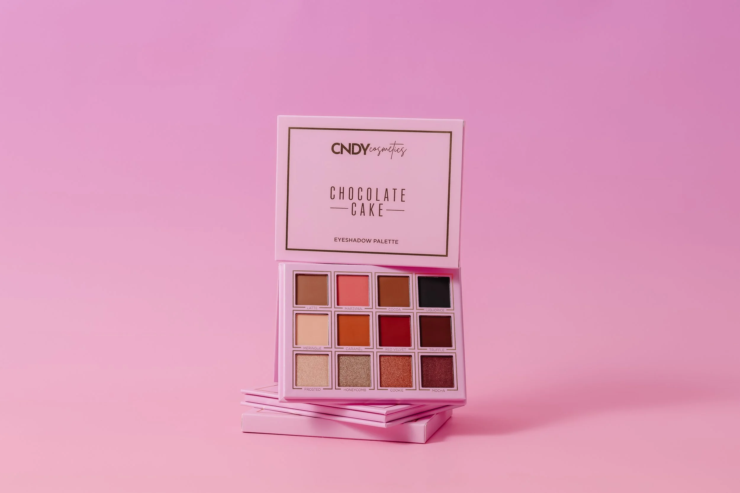 A pink eyeshadow palette with nine shades labeled as Latte, Marzipan, Cocoa, Licorice, Meringue, Caramel, Red Velvet, Truffle, Frosted, Honeycomb, Cookie, Mocha, arranged in three rows against a pink background.
