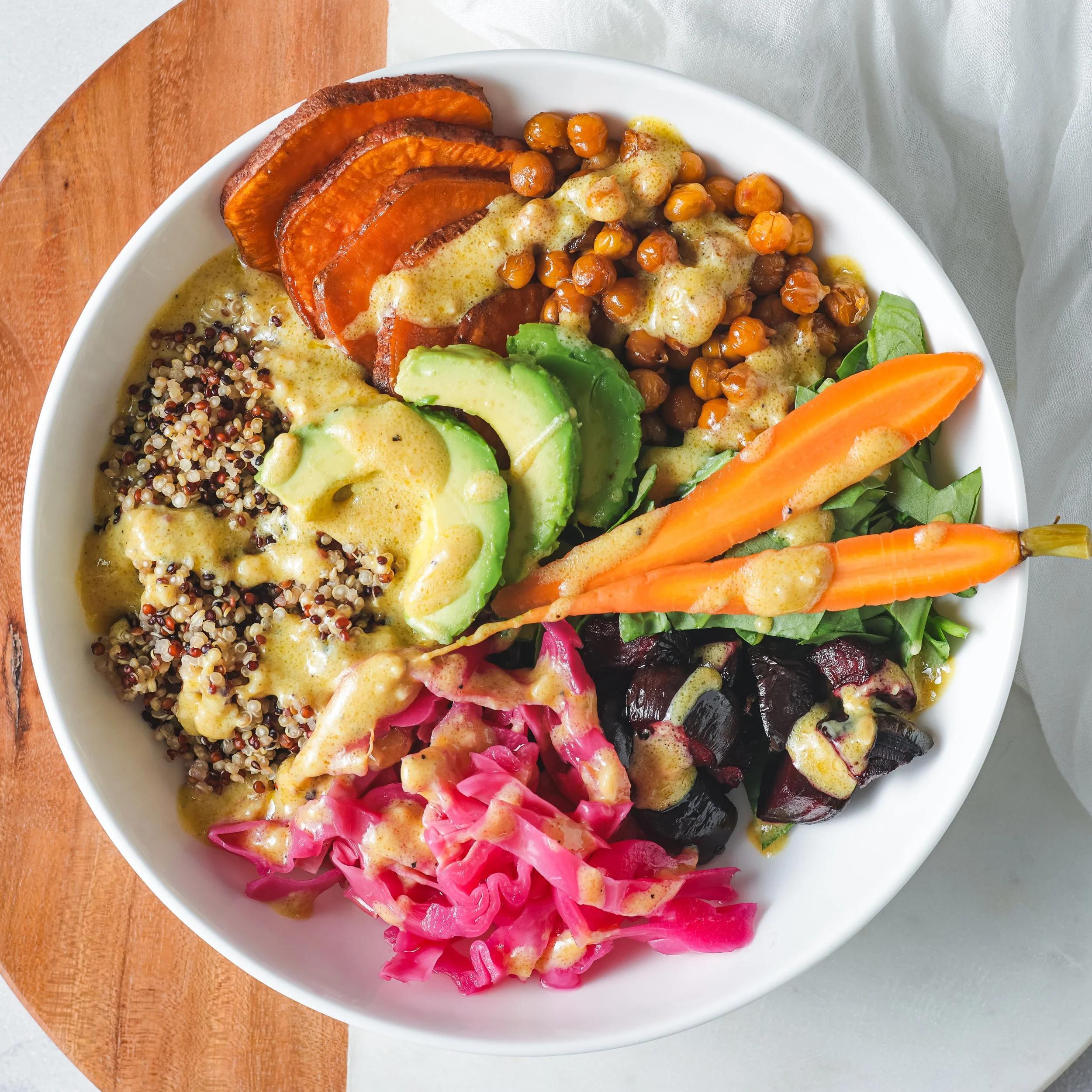 A bowl filled with various colorful foods including roasted pumpkin slices, a mixture of quinoa, chickpeas with sauce, sliced avocado, carrots, beets, and fresh greens on top.
