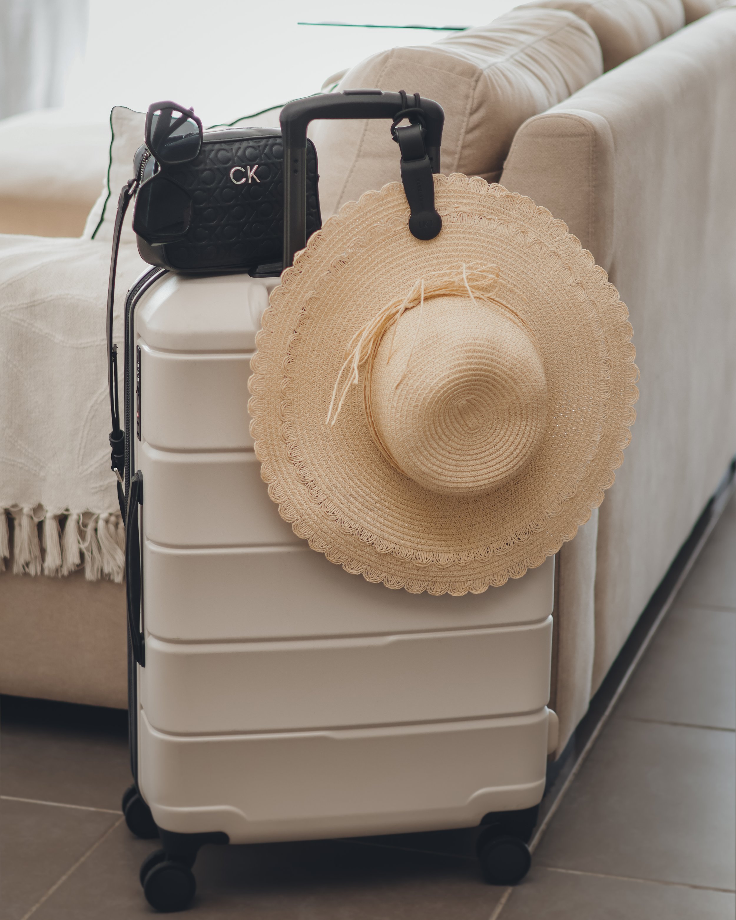 A white rolling suitcase with four black wheels, a black CK handbag, a beige wide-brimmed straw hat, and a black pair of sunglasses resting on the suitcase, all placed beside a beige sofa with tufted cushions and a throw pillow.