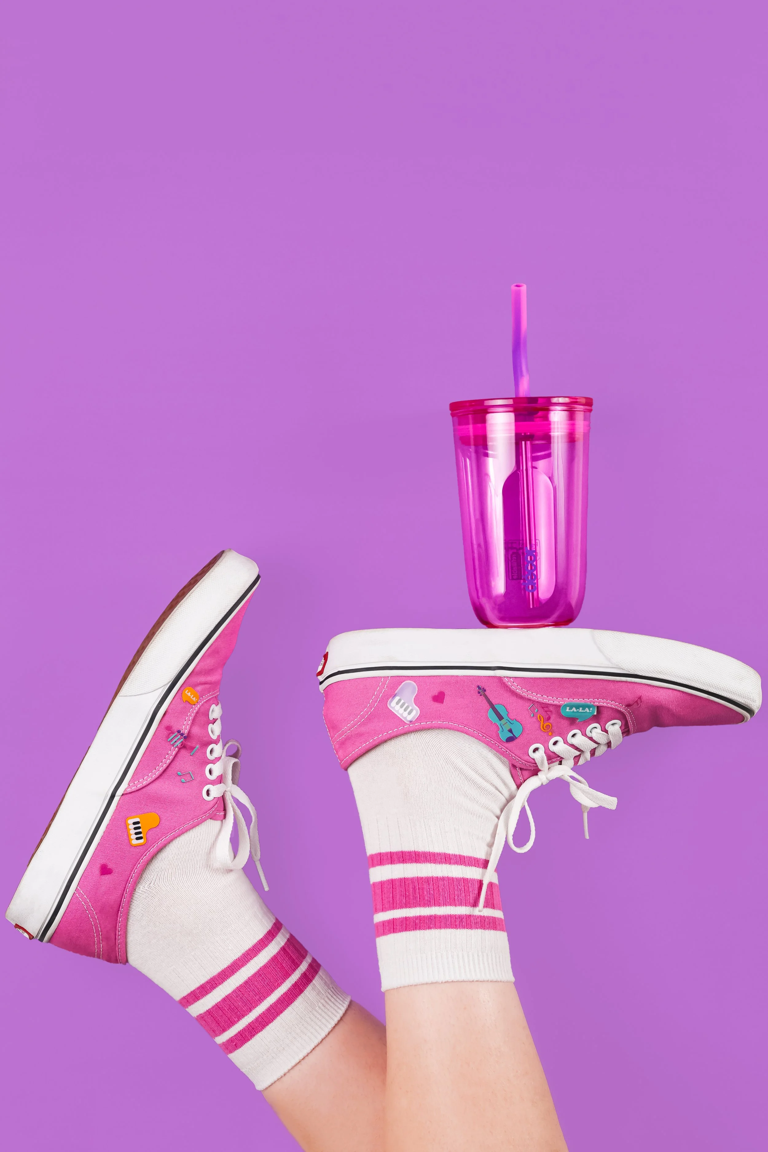 Person wearing pink and white sneakers with music, heart, and guitar patches, striped pink and white socks, holding a pink cup with straw against a purple background.