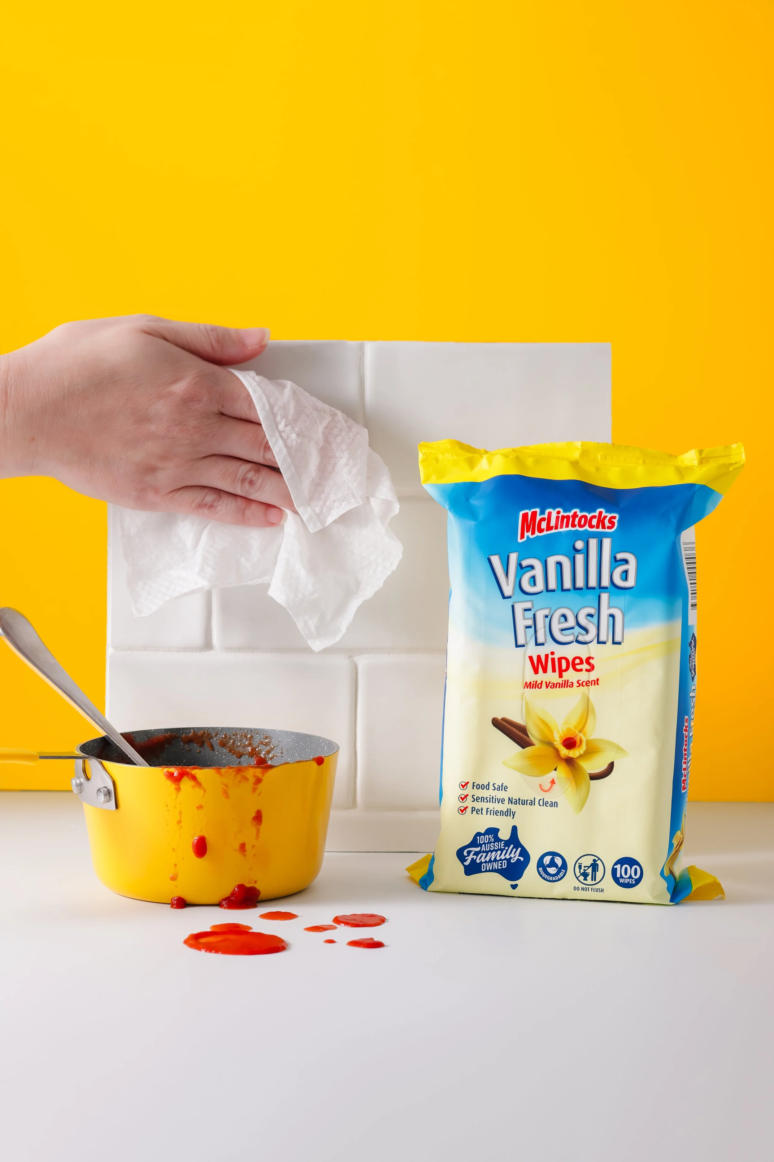 A hand wiping the white tiled wall behind a bag of McIntocks Vanilla Fresh Wipes. A yellow baking dish with spilled red sauce and a spoon is on the table, with the sauce dripping down the side. The background is yellow with white tiles.