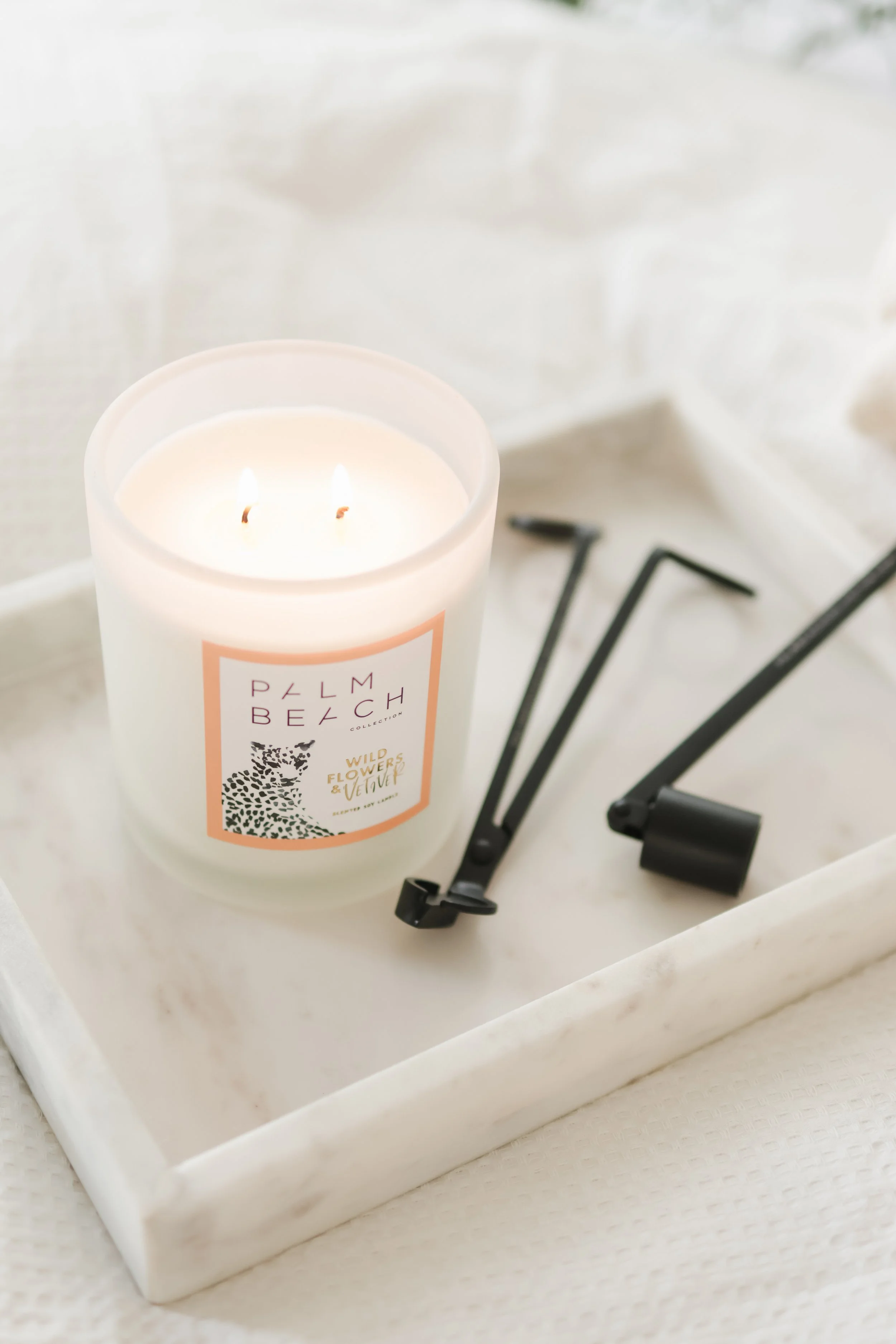 A lit white candle with a label reading 'Palm Beach Collection Wild Flowers & Vetiver' is placed on a white tray alongside black and silver eyelash curlers, on a white surface.