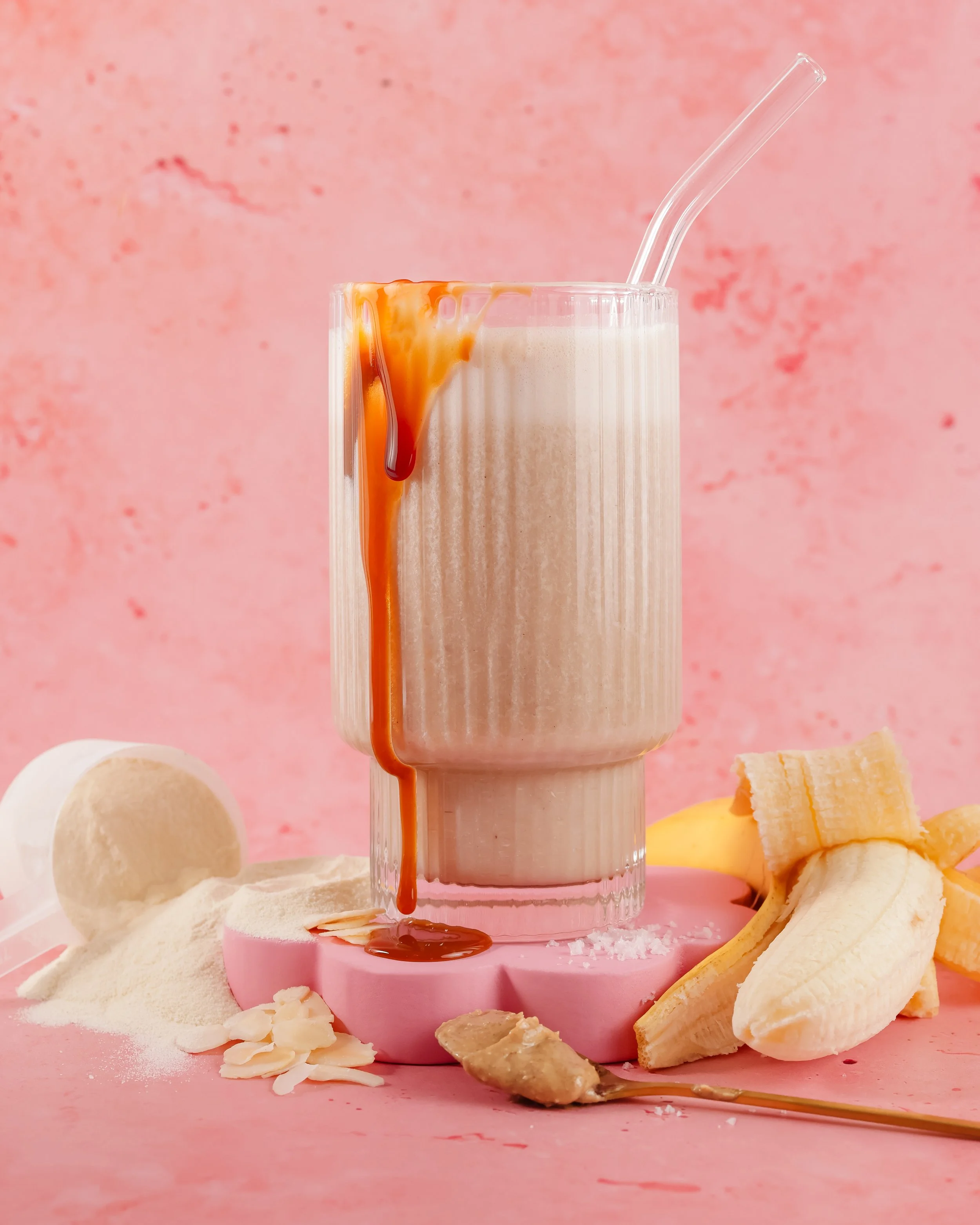 A glass of banana-flavored milkshake with caramel sauce dripping over the side, set against a pink background. Sliced bananas and ice cream are positioned around the glass.