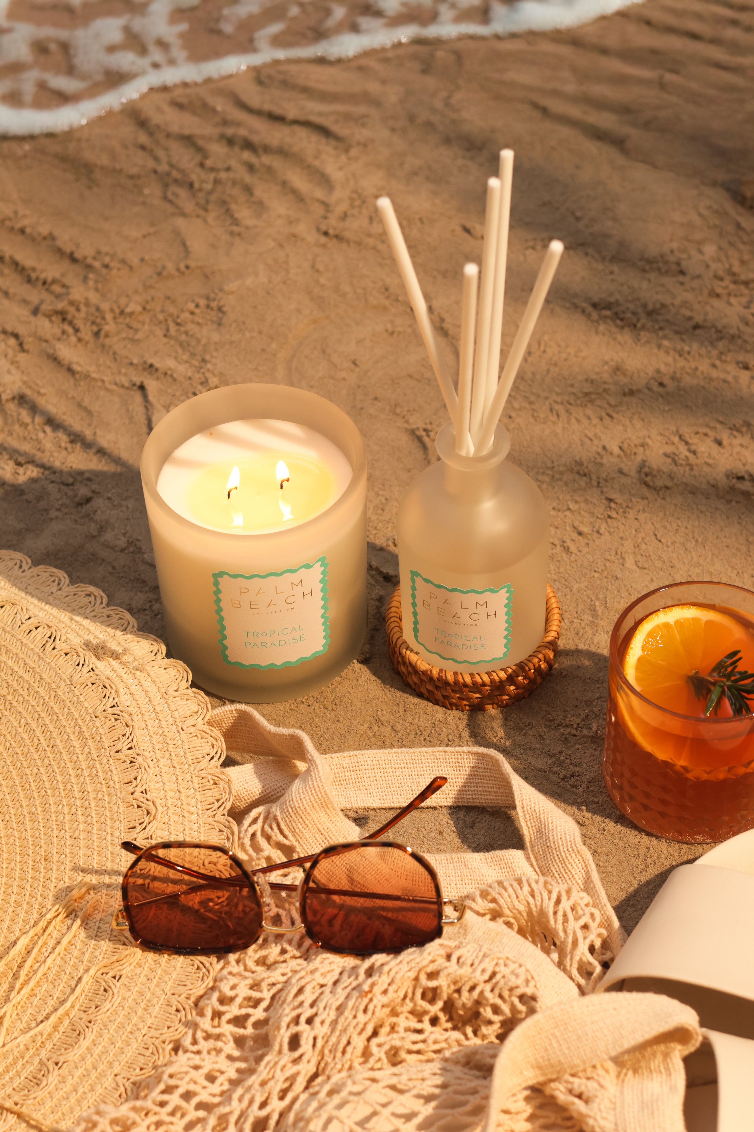 Beachside scene featuring a lit candle, reed diffuser, sunglasses, a plate with orange slices, and a crocheted cover-up on the sand.