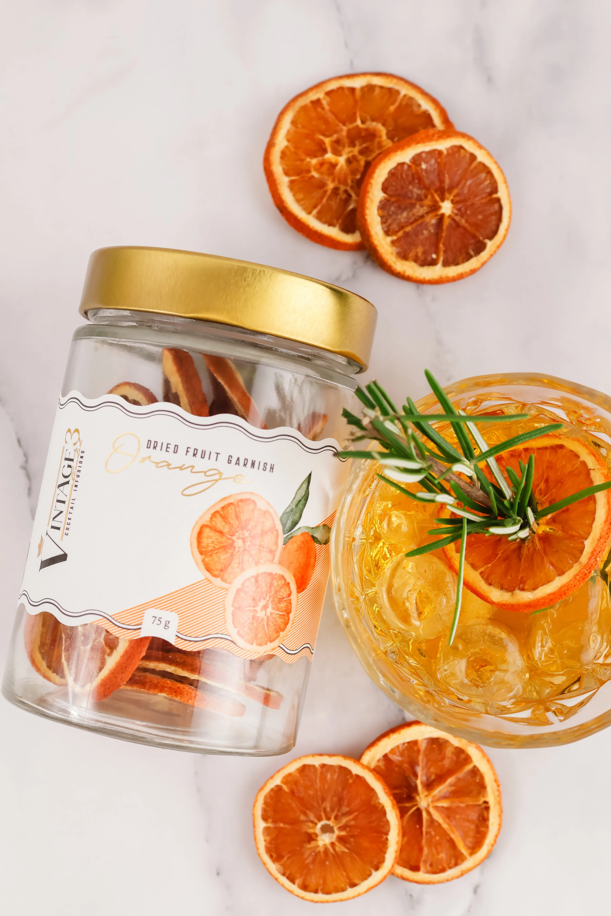 A jar of dried orange fruit garnish with a label and a glass bowl filled with ice, orange slices, and sprigs of rosemary on a white marble surface.