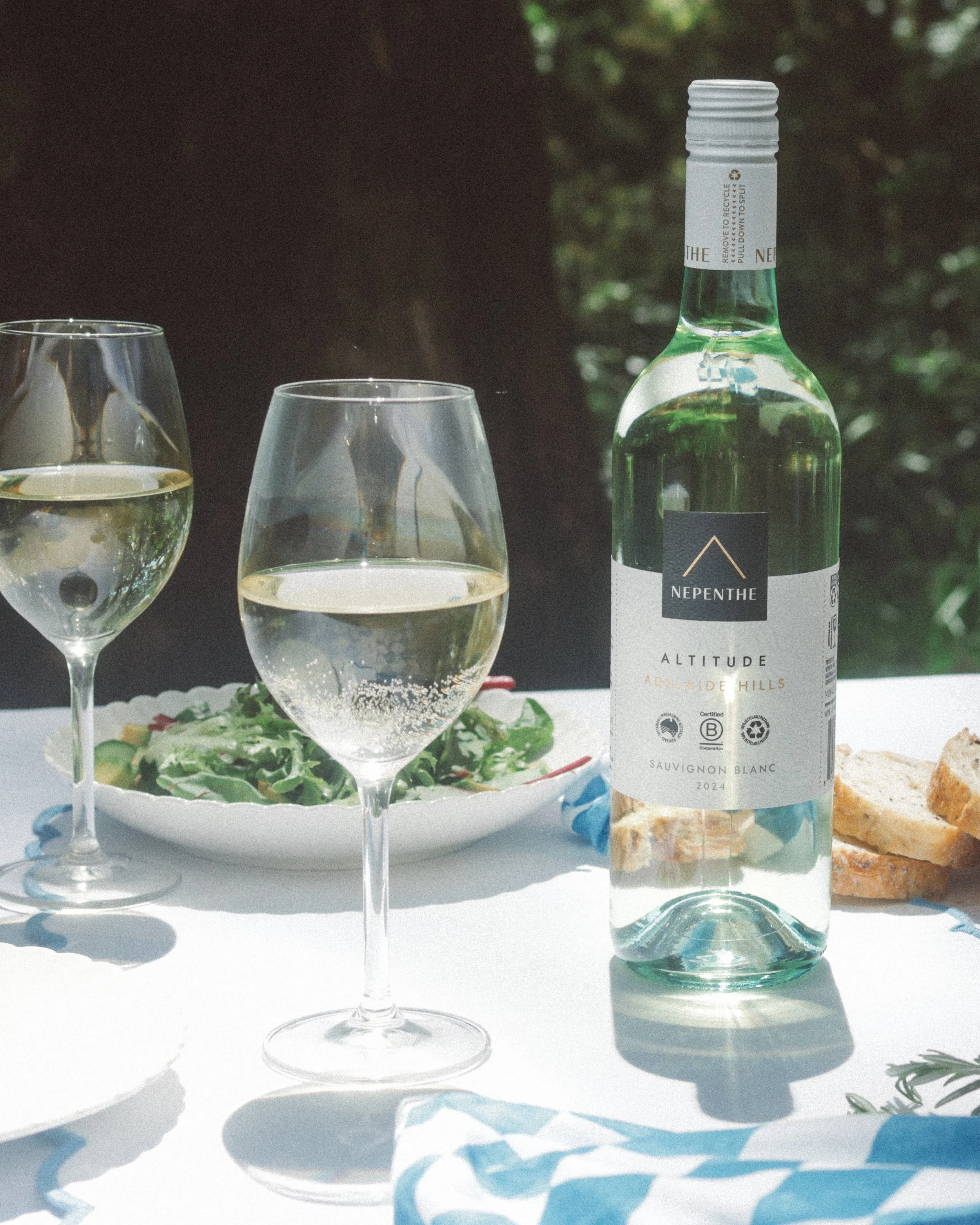A bottle of Nepenthe Altitude Sauvignon Blanc white wine with two wine glasses filled with white wine, a salad with greens and red peppers on a white plate, and some bread on a table outdoors with green foliage in the background.