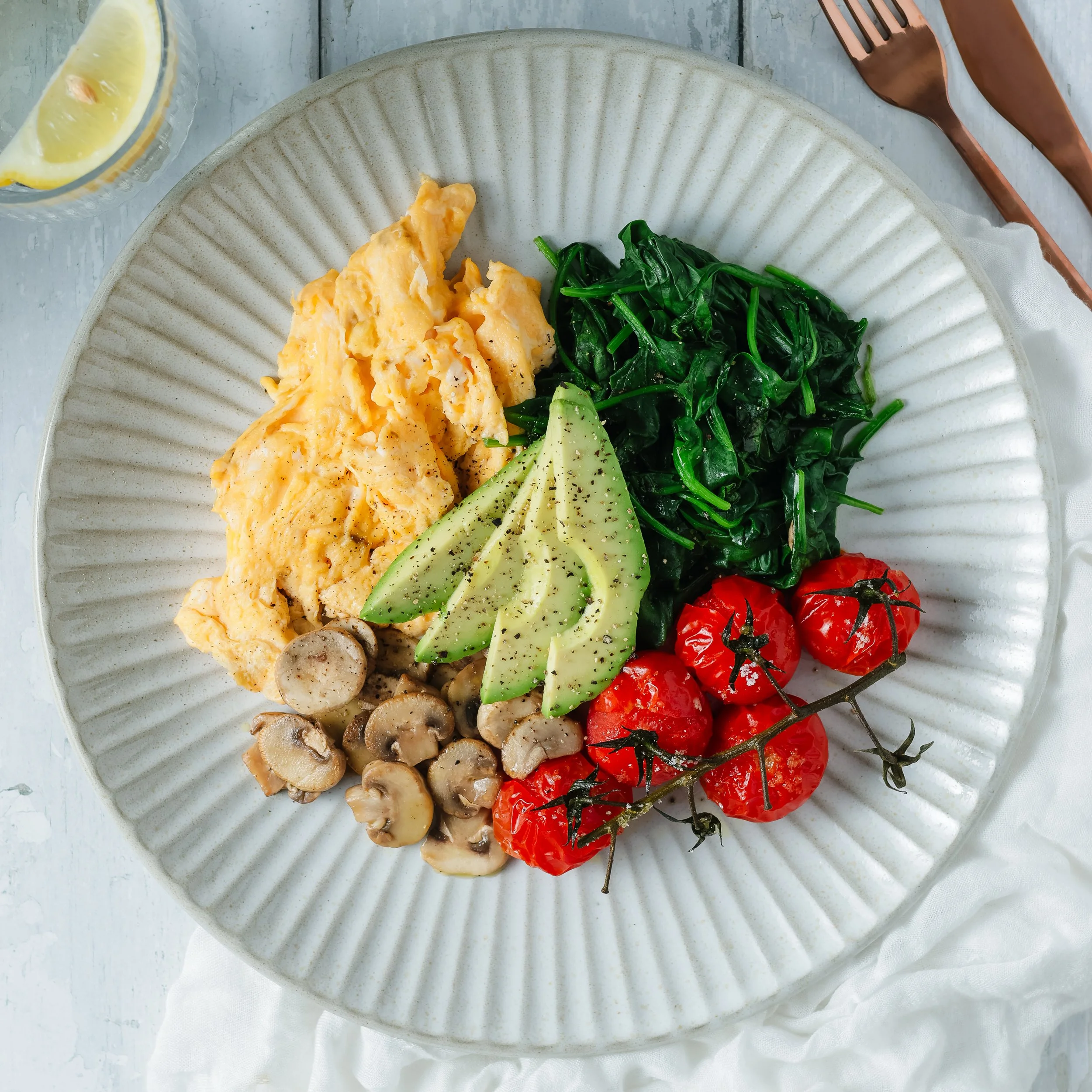 A white ceramic plate with scrambled eggs, sautéed spinach, sliced cucumbers, cherry tomatoes, and mushrooms. A glass of lemon water is partially visible at the top left, with a copper-colored fork and knife set on the right side, and a white cloth n