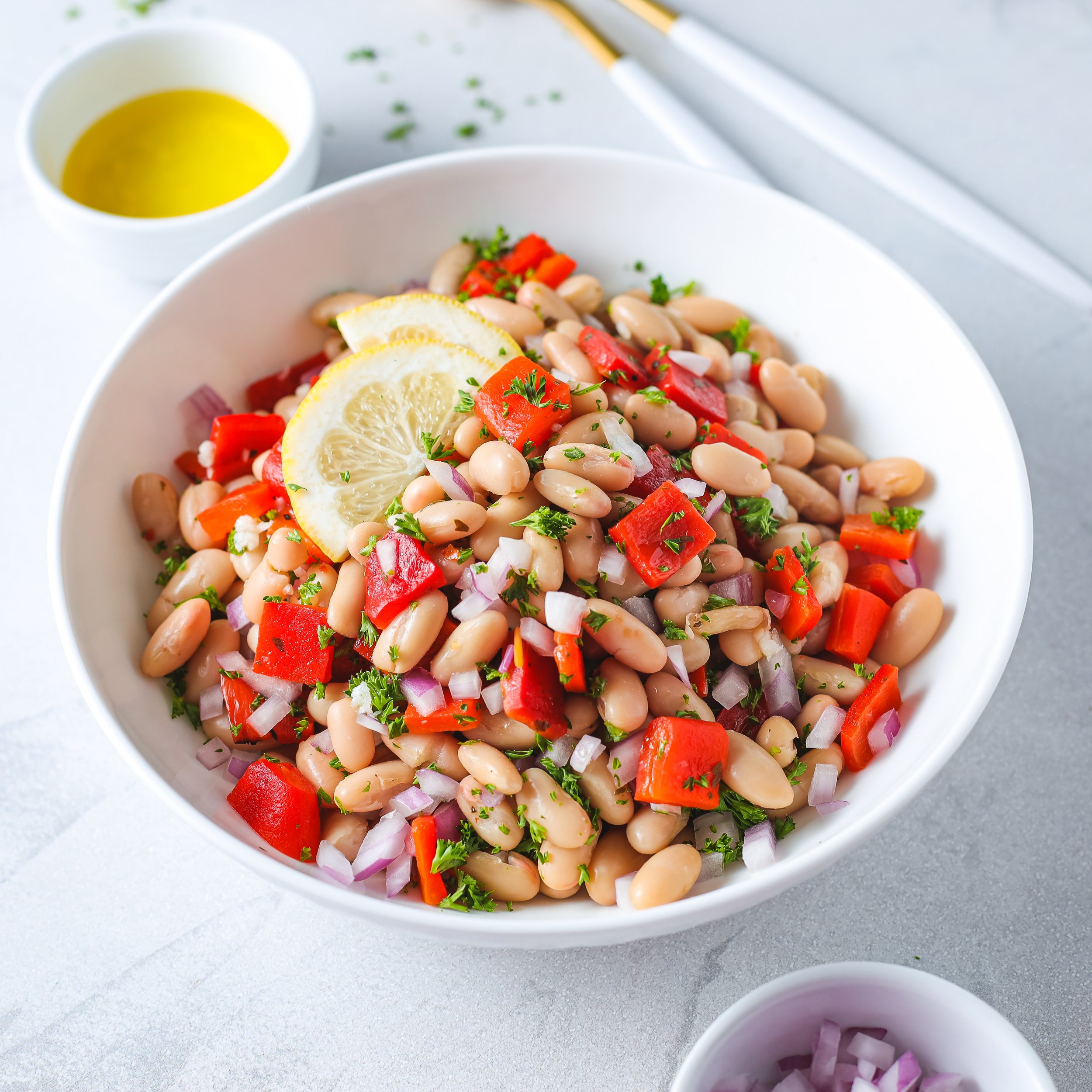 A white bowl of bean salad garnished with lemon slices and chopped parsley, accompanied by small bowls of olive oil and chopped red onions.