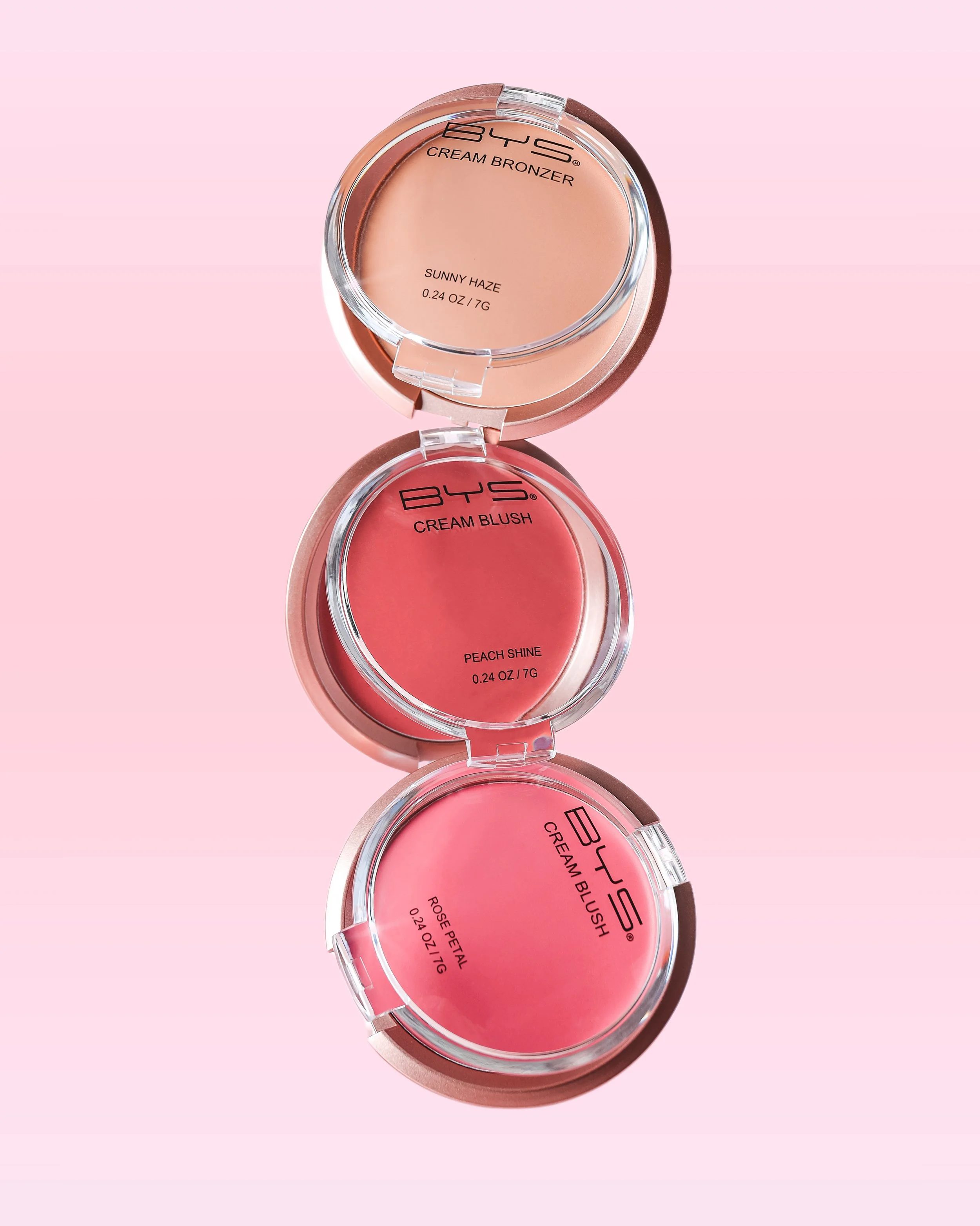 Three round compact makeup containers stacked vertically on a pink background. The top container labeled 'BYS Cream Bronzer' in shade 'Sunny Haze,' the middle labeled 'BYS Cream Blush' in shade 'Peach Shine,' and the bottom container labeled 'BYS Cre