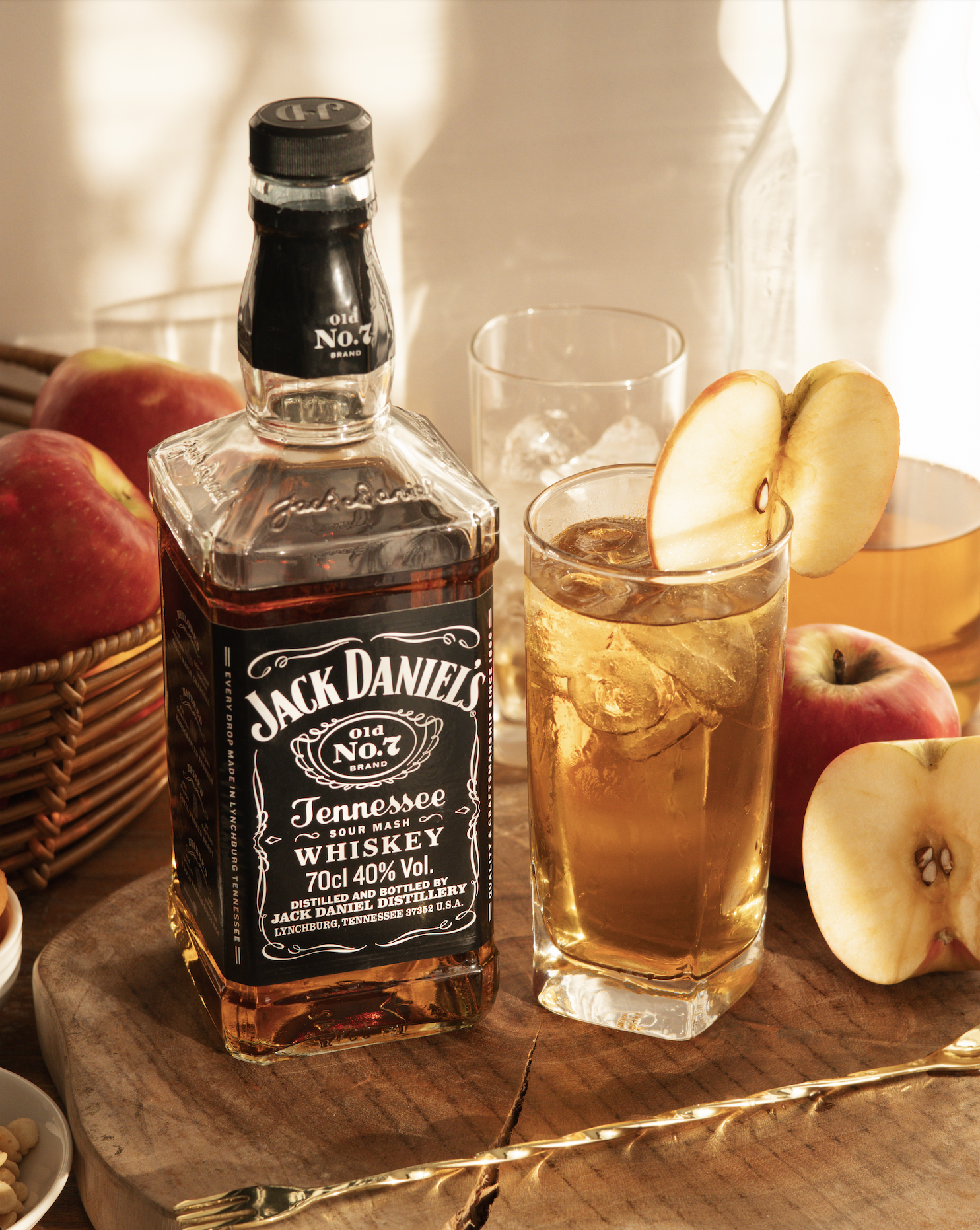 A bottle of Jack Daniel's Tennessee whiskey, a glass of whiskey with ice and a slice of apple, round red apples, a glass with ice, a white pitcher, a basket of apples, a small dish of peanuts, and a gold fork on a wooden table.