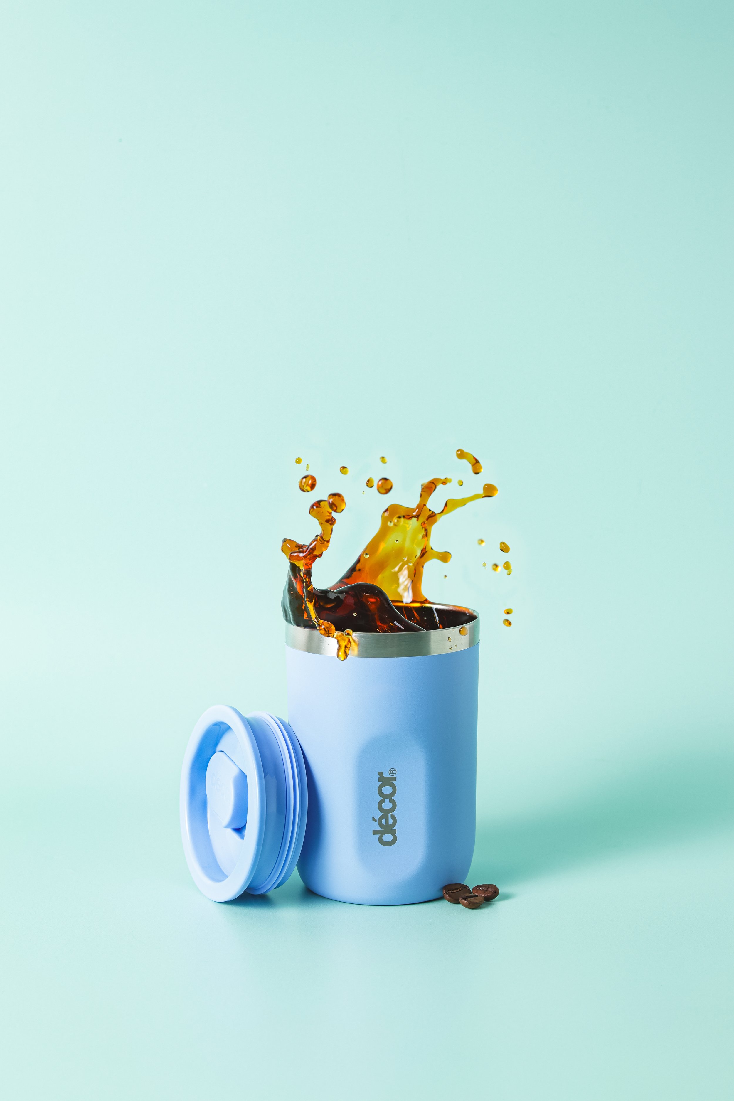 A blue coffee capsule with its lid off, spilling coffee into a small cup with coffee beans nearby, on a mint green background.