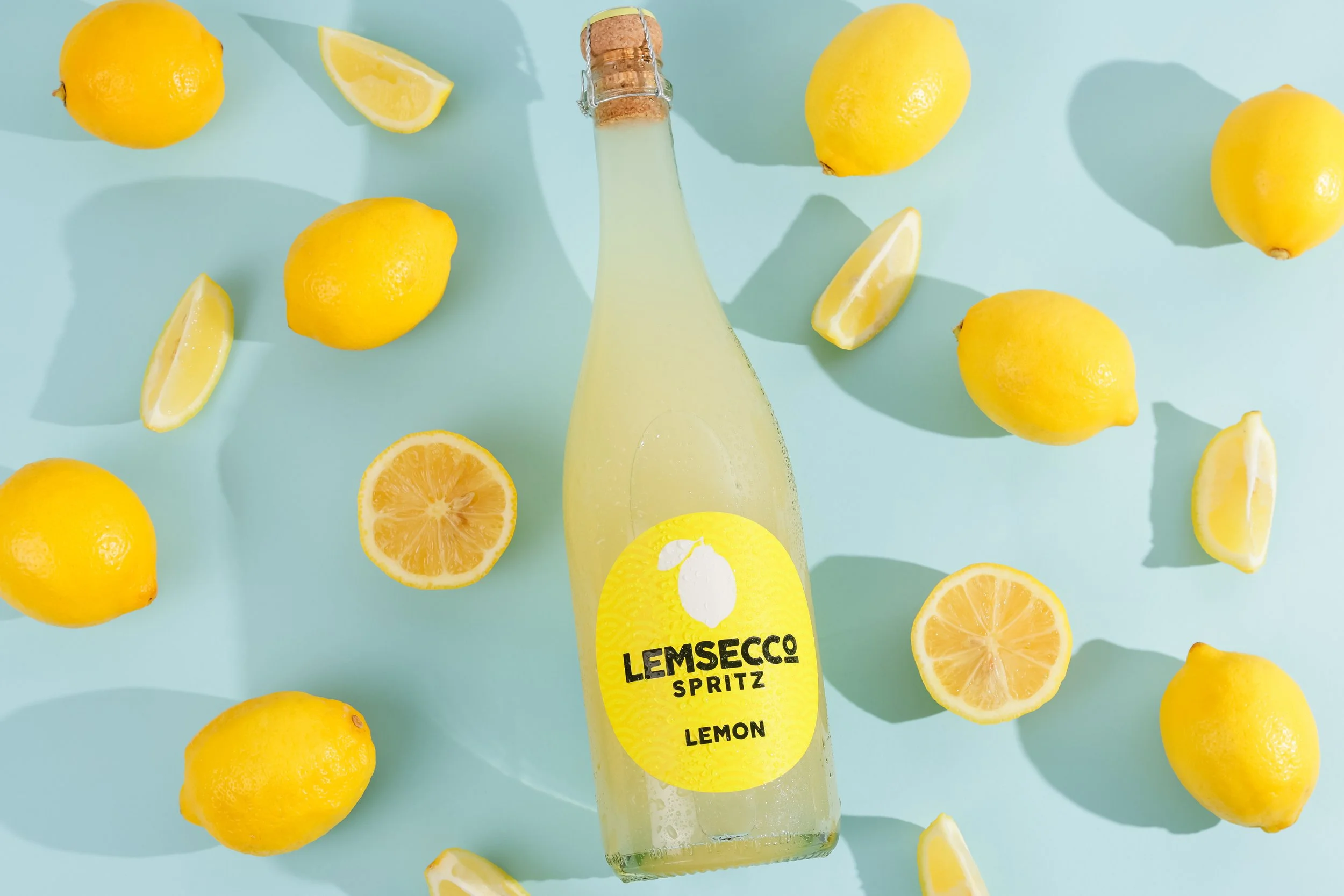 A glass bottle labeled 'Lemsecco Spritz Lemon' surrounded by whole and sliced lemons on a light blue surface.