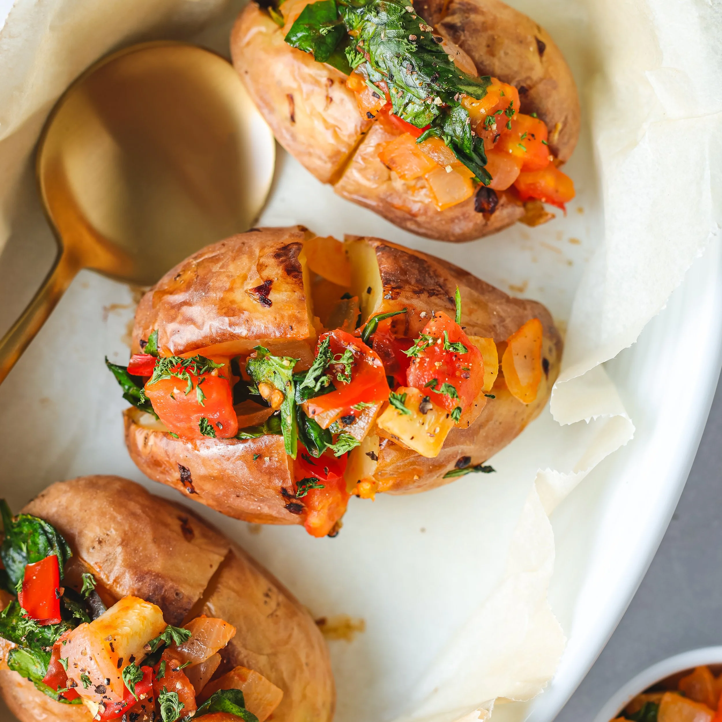 Close-up of baked potatoes topped with tomato, green herbs, and seasonings, served with a side of creamy sauce.