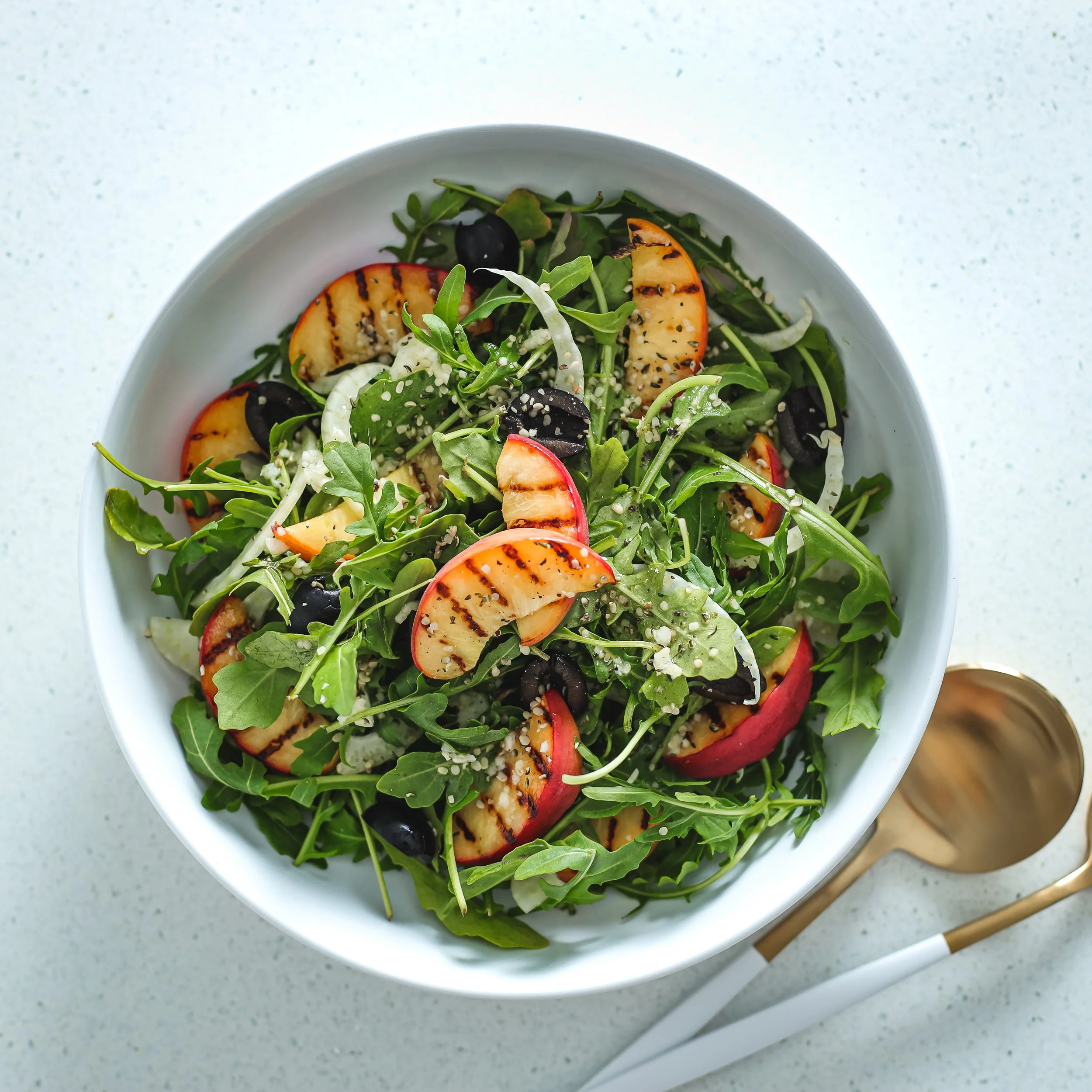A fresh salad in a white bowl containing grilled peach slices, arugula, black olives, shredded cheese, and sesame seeds on top.