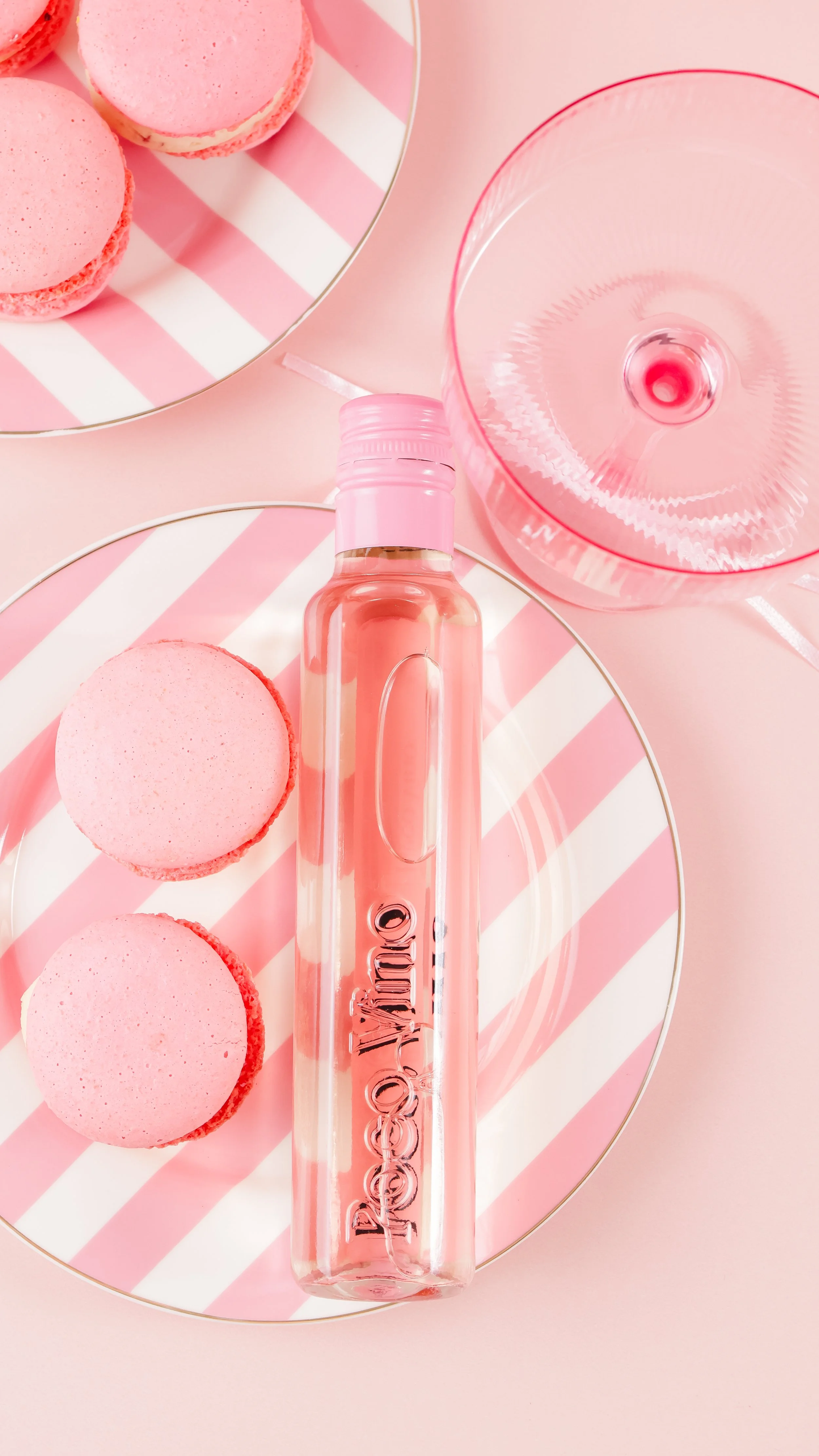 Pink macarons on a striped pink and white plate, a pink glass, and a clear pink bottle with the word 'CHROME' on it, all arranged on a pink background.