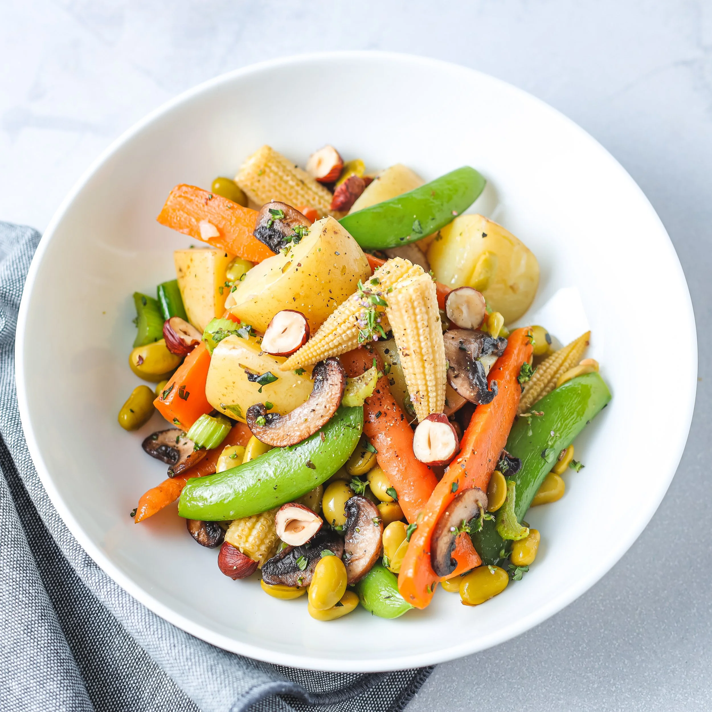 A white bowl filled with a mixed vegetable salad including carrots, snap peas, baby corn, potatoes, mushrooms, and herbs, sitting on a gray surface with a gray cloth underneath.