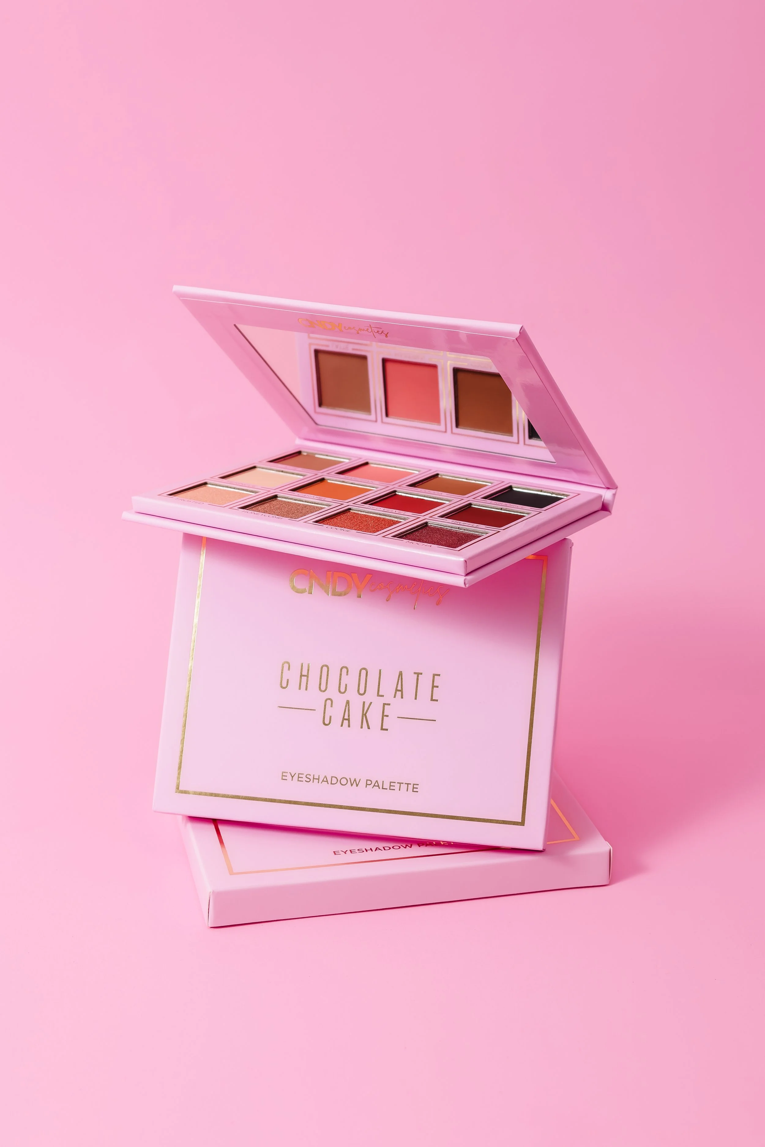 Open pink eyeshadow palette with 12 shades displayed on top of two pink boxes against pink background.