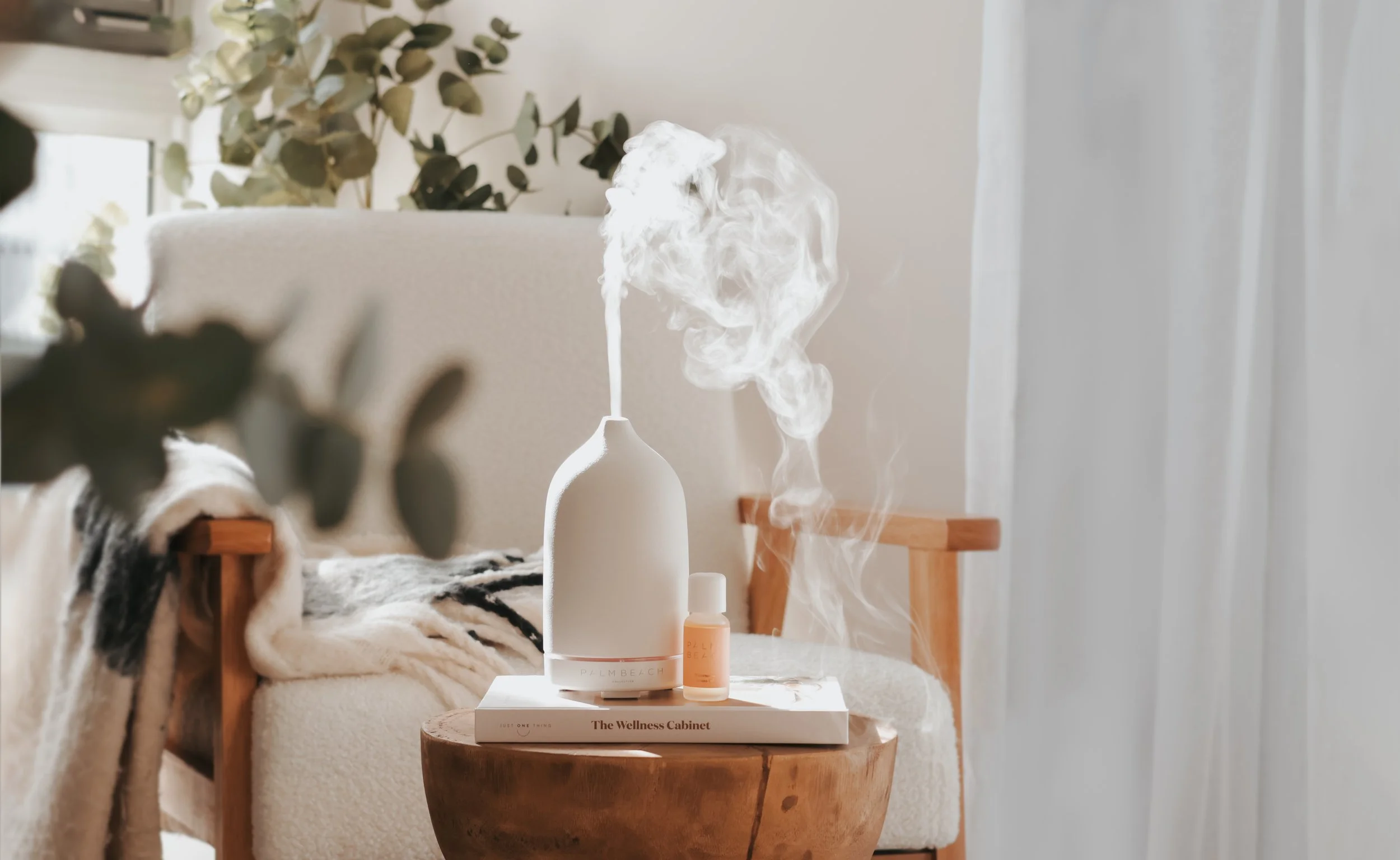 A bedside table with a white diffuser emitting steam, a small bottle, and a stack of books, including one titled "The Wellness Cabinet," in a cozy room with a beige couch and greenery in the background.