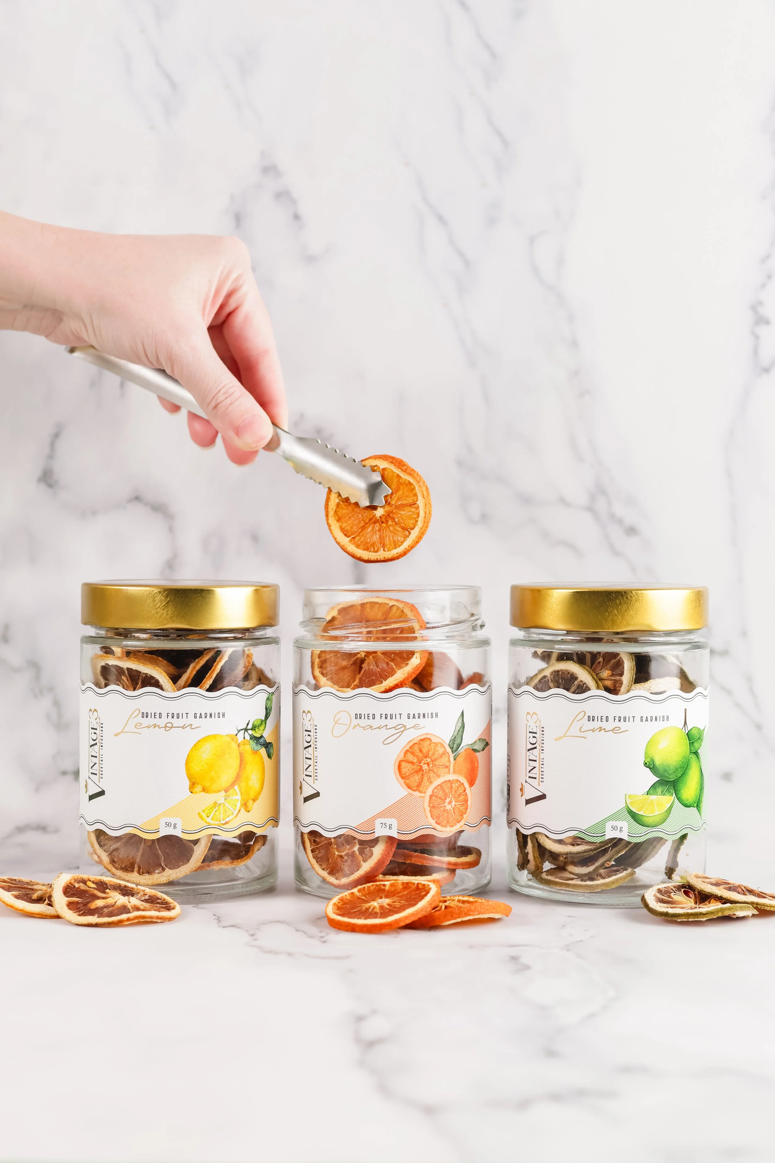 Three glass jars filled with dried fruit slices, labeled as lemon, orange, and lime flavored dried fruit garnishes, sitting on a white marble surface with scattered dried fruit slices in front. A hand is holding tongs with a dried orange slice, about