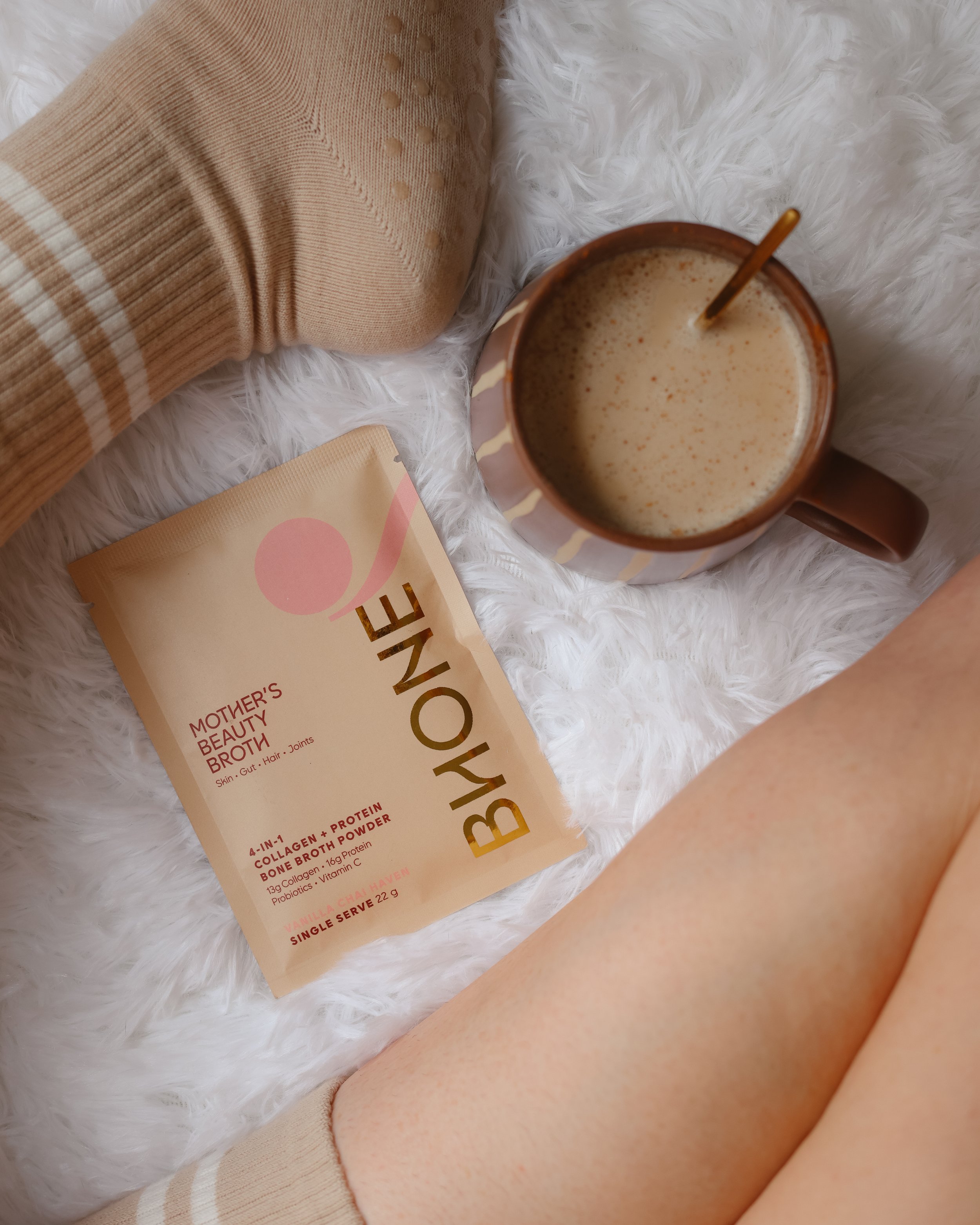 Close-up of a beige sock, a white fluffy surface, a tan mug with a drink inside, a beige packet of collagen bone broth powder, and part of a person's leg.