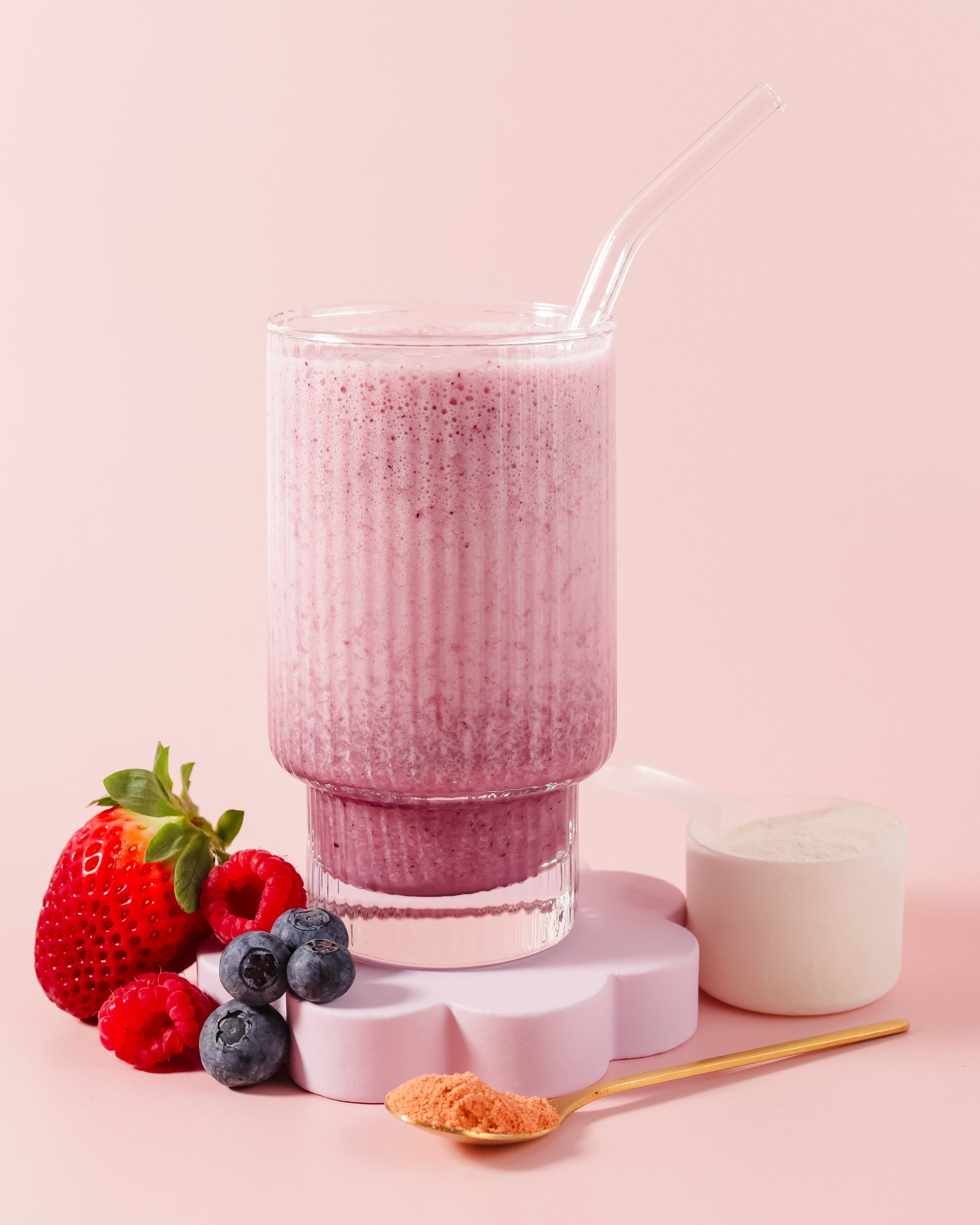 A glass of pink strawberry smoothie with a clear straw, surrounded by fresh strawberries, blueberries, raspberries, and a dollop of cream on a pink background, with a gold spoon containing pink powder and white dessert disks.